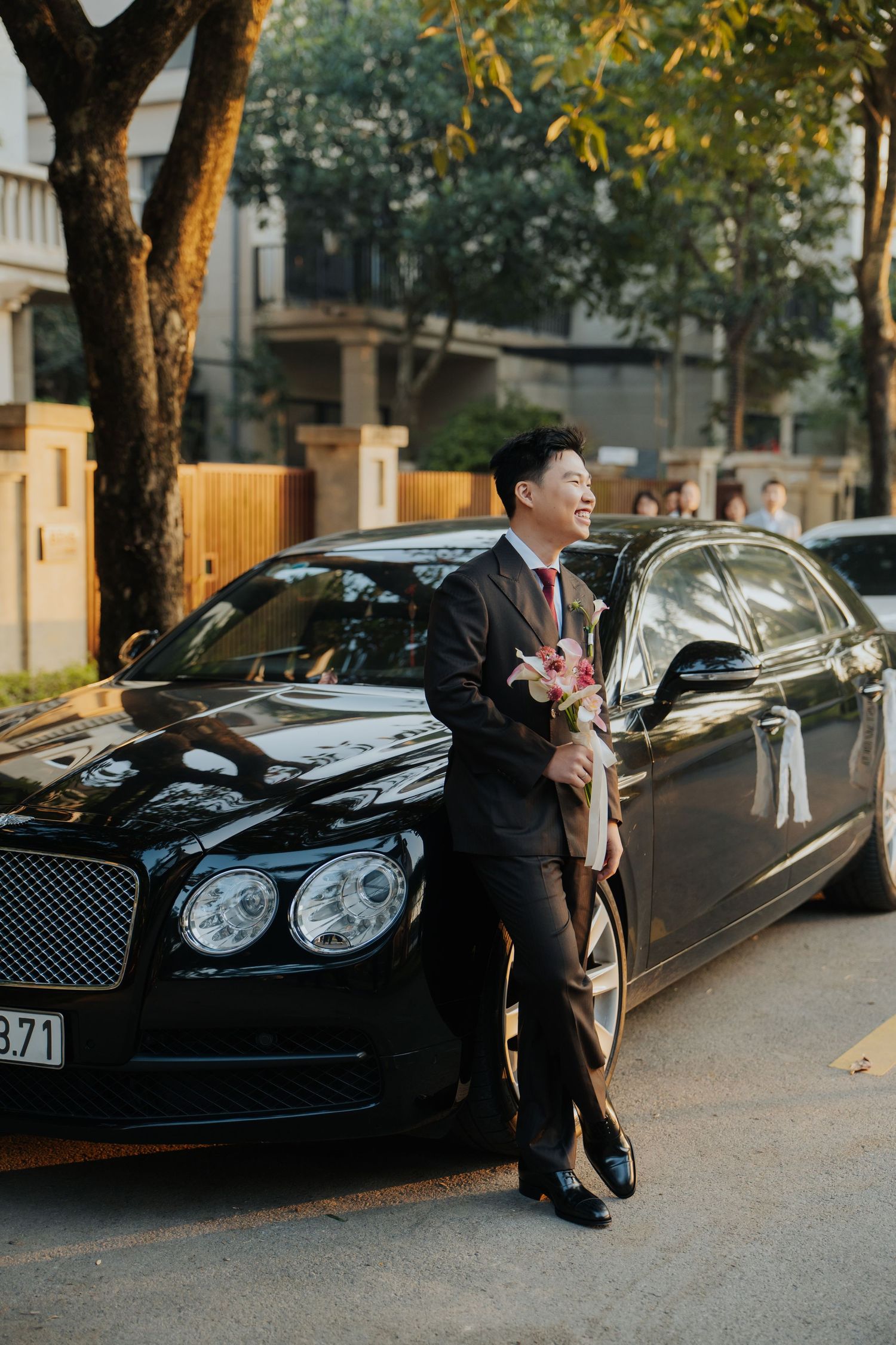 Smiling groom in dark suit holding bouquet leaning against black Bentley luxury car on street.