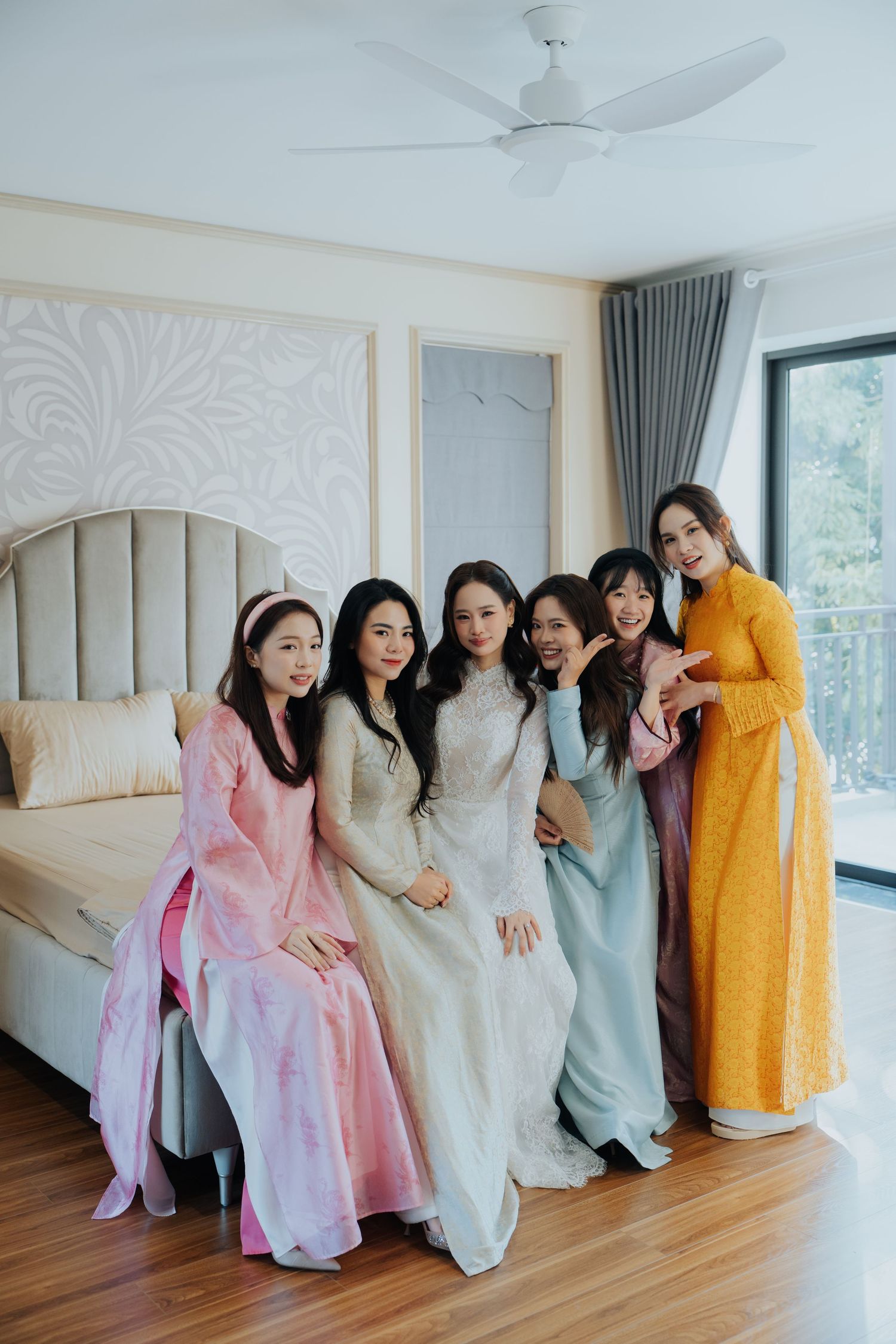 Six women in colorful Vietnamese ao dai dresses pose together in an elegant bedroom, smiling for a group photo.