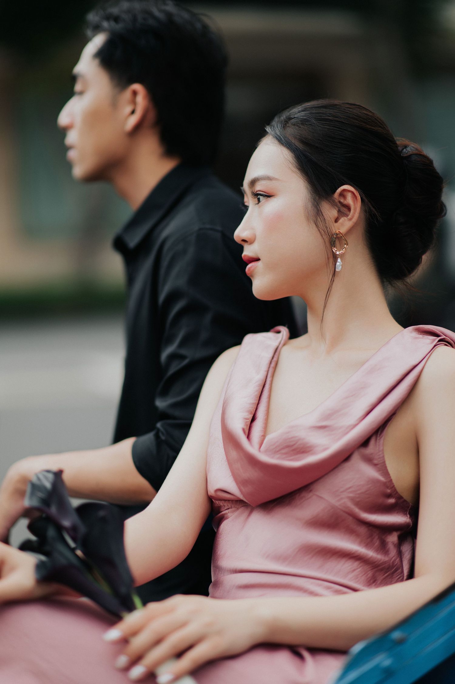 A young Asian woman in a pink satin dress sits beside a man in black, both gazing away in a candid outdoor portrait.