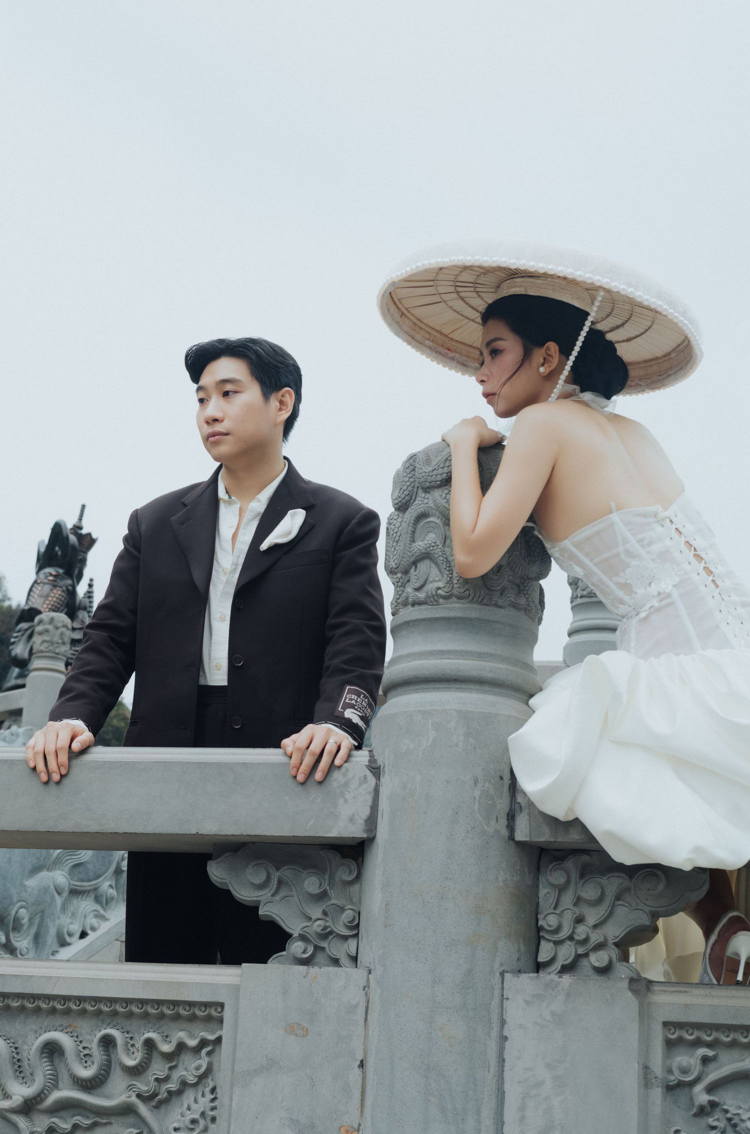 Couple in wedding attire at ornate stone balustrade, groom in dark suit, bride in white dress with traditional Asian hat.