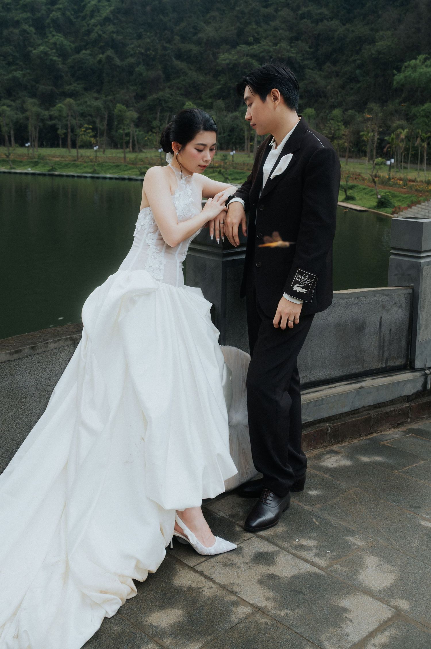 A bride in a white gown and groom in a black suit pose romantically by a lakeside stone railing with lush greenery.