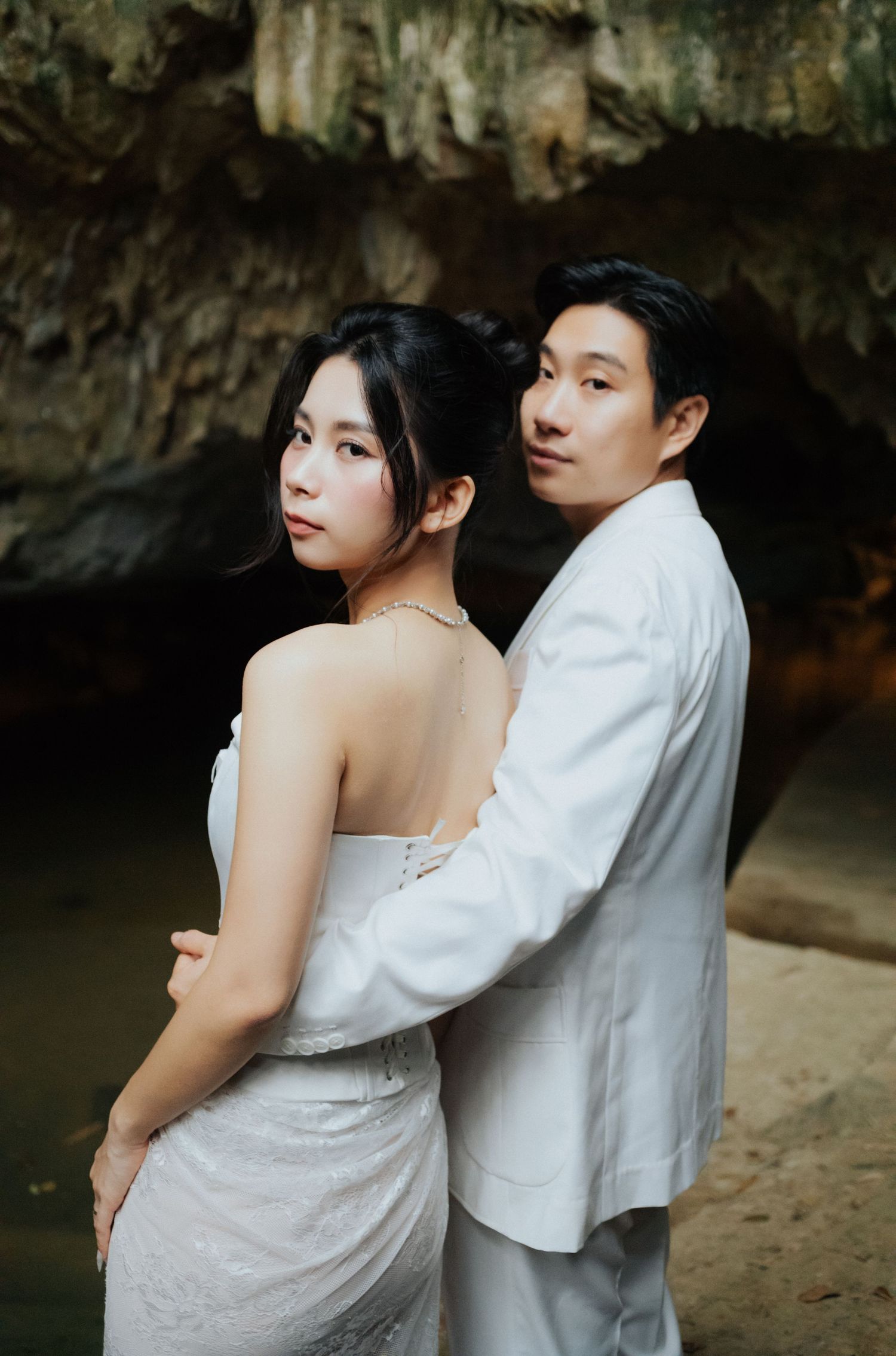 Couple in white attire posing romantically in a cave, woman in backless dress looking over shoulder.