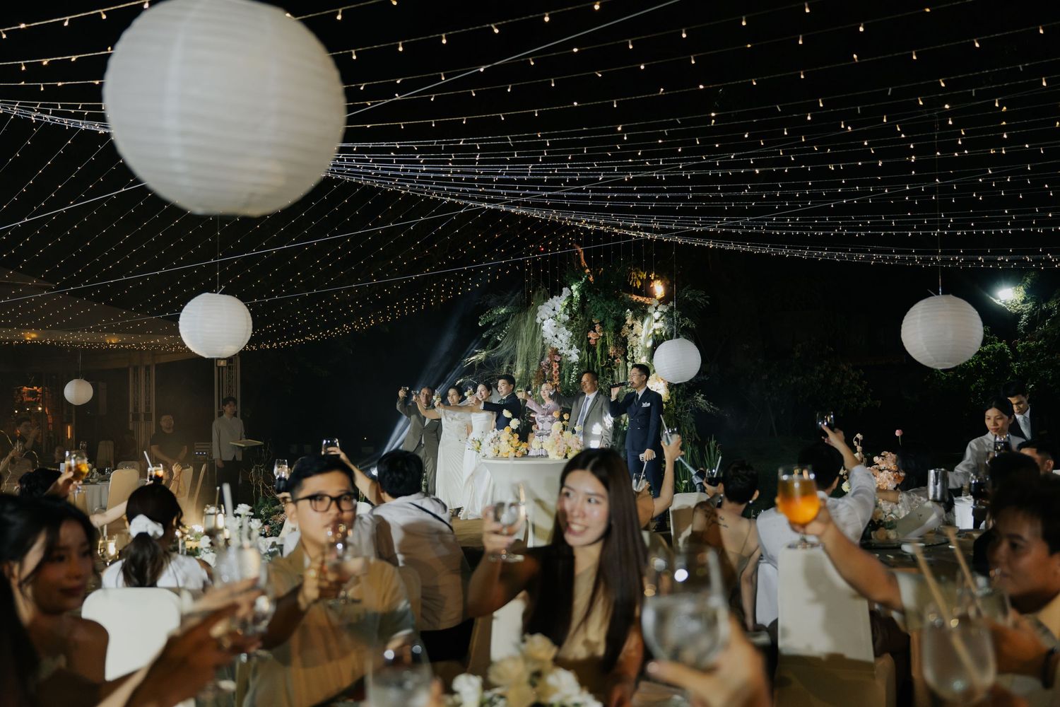 Elegant outdoor wedding reception at night with string lights, white lanterns, and guests celebrating at decorated tables.
