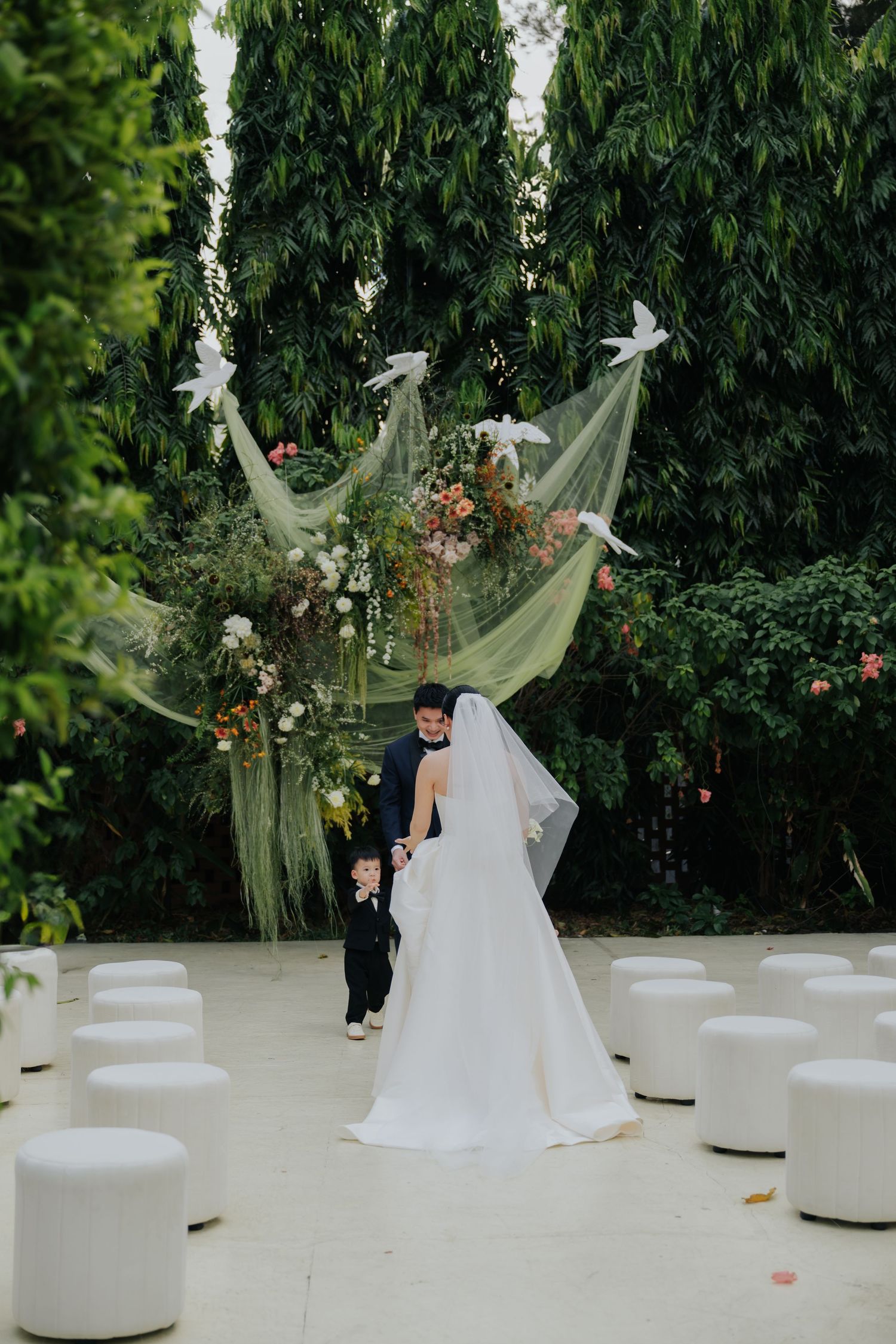 Bride and groom stand with young child at outdoor wedding ceremony altar adorned with greenery, flowers, and white doves.