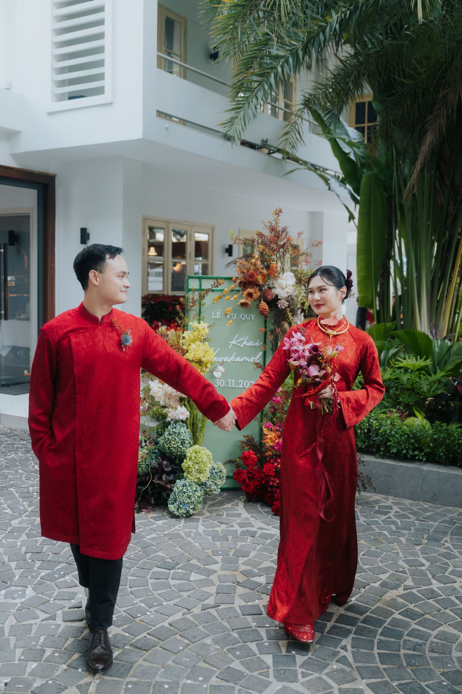 Vietnamese couple in red ao dai traditional wedding attire holding hands in a lush garden courtyard.