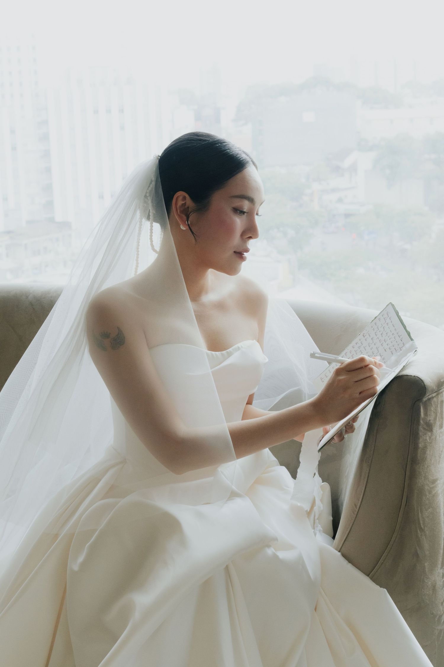 A bride in a white strapless gown and veil sits in a chair by a window, writing in a notebook.
