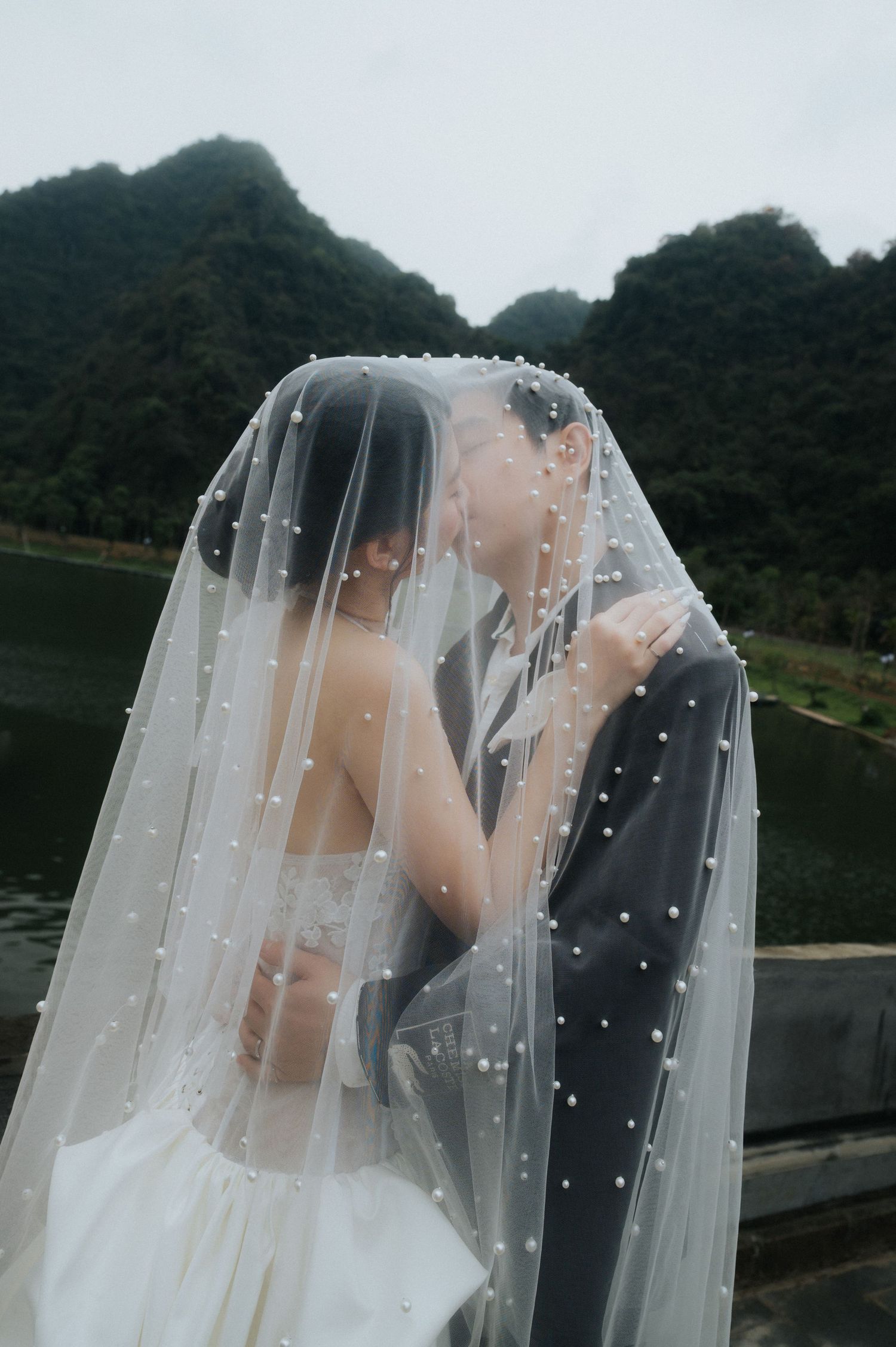 A bride and groom share a romantic kiss under a pearl-adorned wedding veil with misty mountains and a lake in the background.