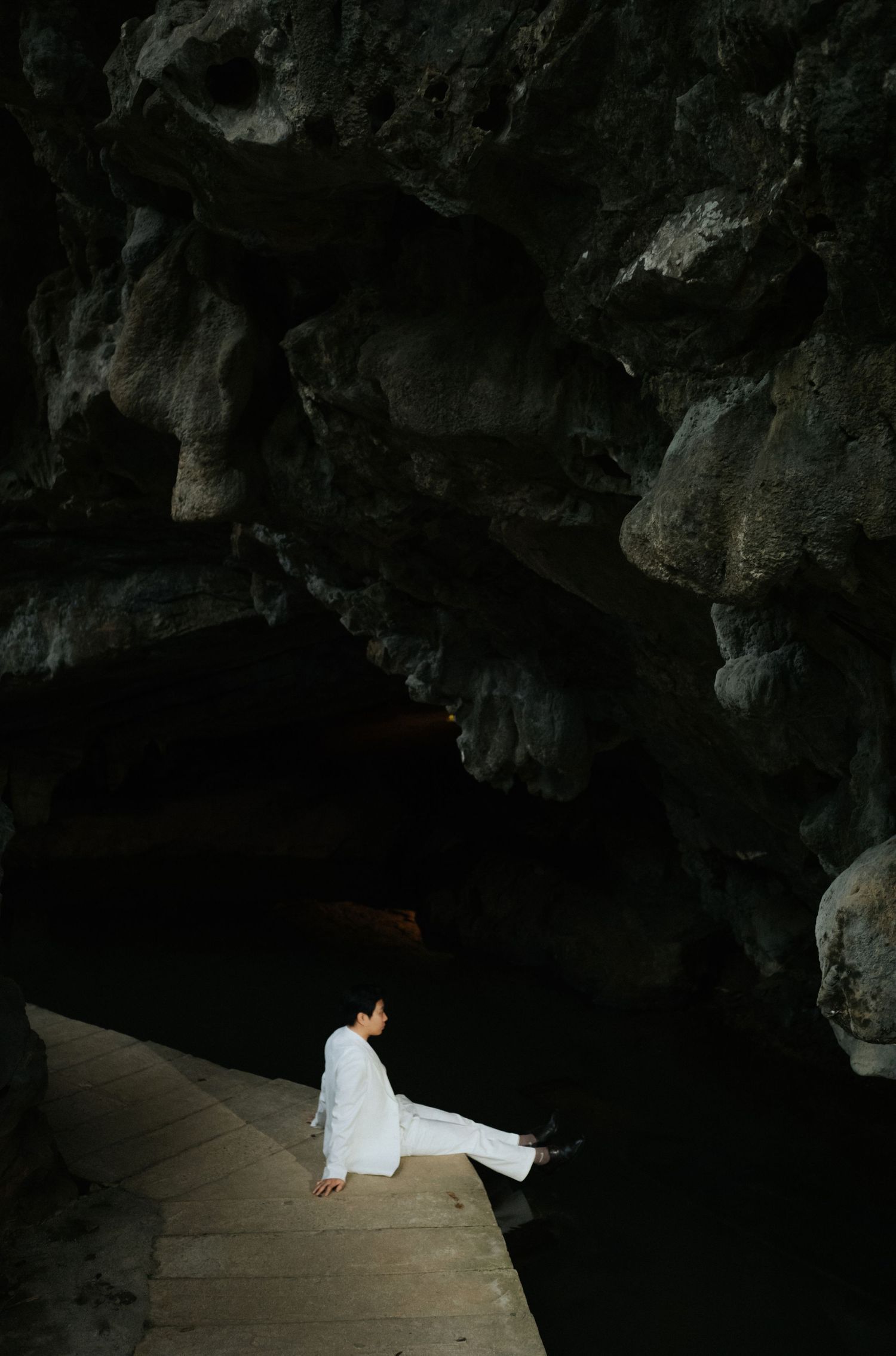 Person in white clothing sitting on wooden steps inside a dark cave with rocky walls and still water.