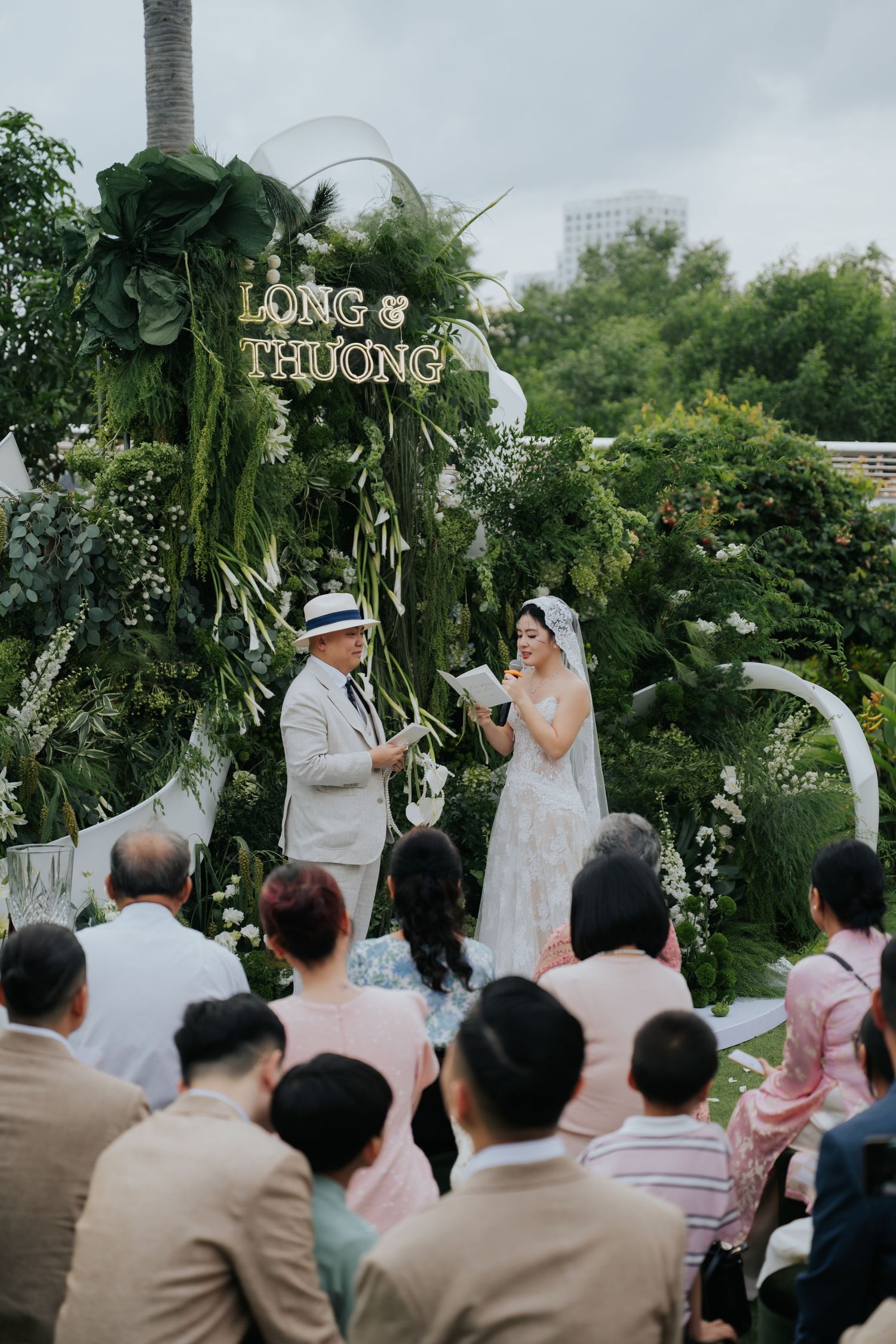 Outdoor wedding ceremony for Long and Thuong, bride in white gown reads vows before seated guests amid lush green floral backdrop.