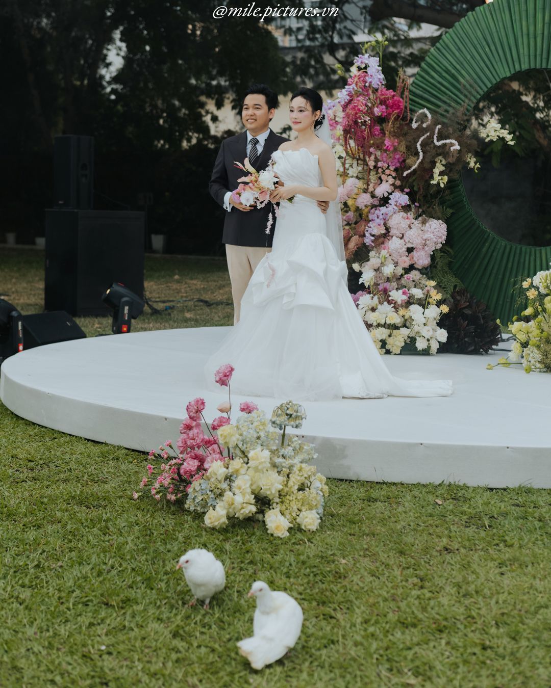 Bride in white gown and groom stand on circular stage with floral backdrop, two white doves on grass below.