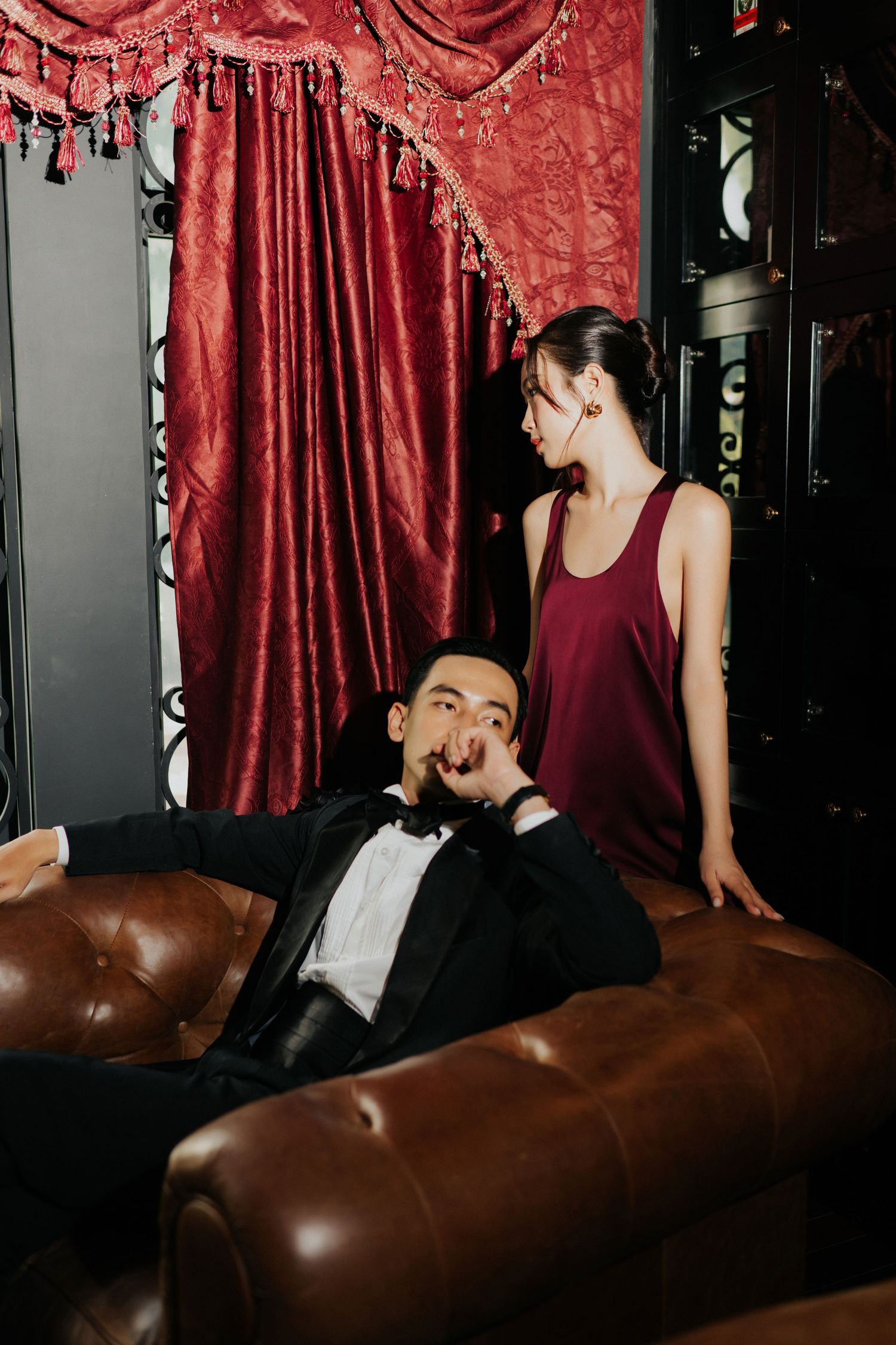 Man in tuxedo reclines on leather sofa beside woman in burgundy dress, red velvet curtains backdrop.