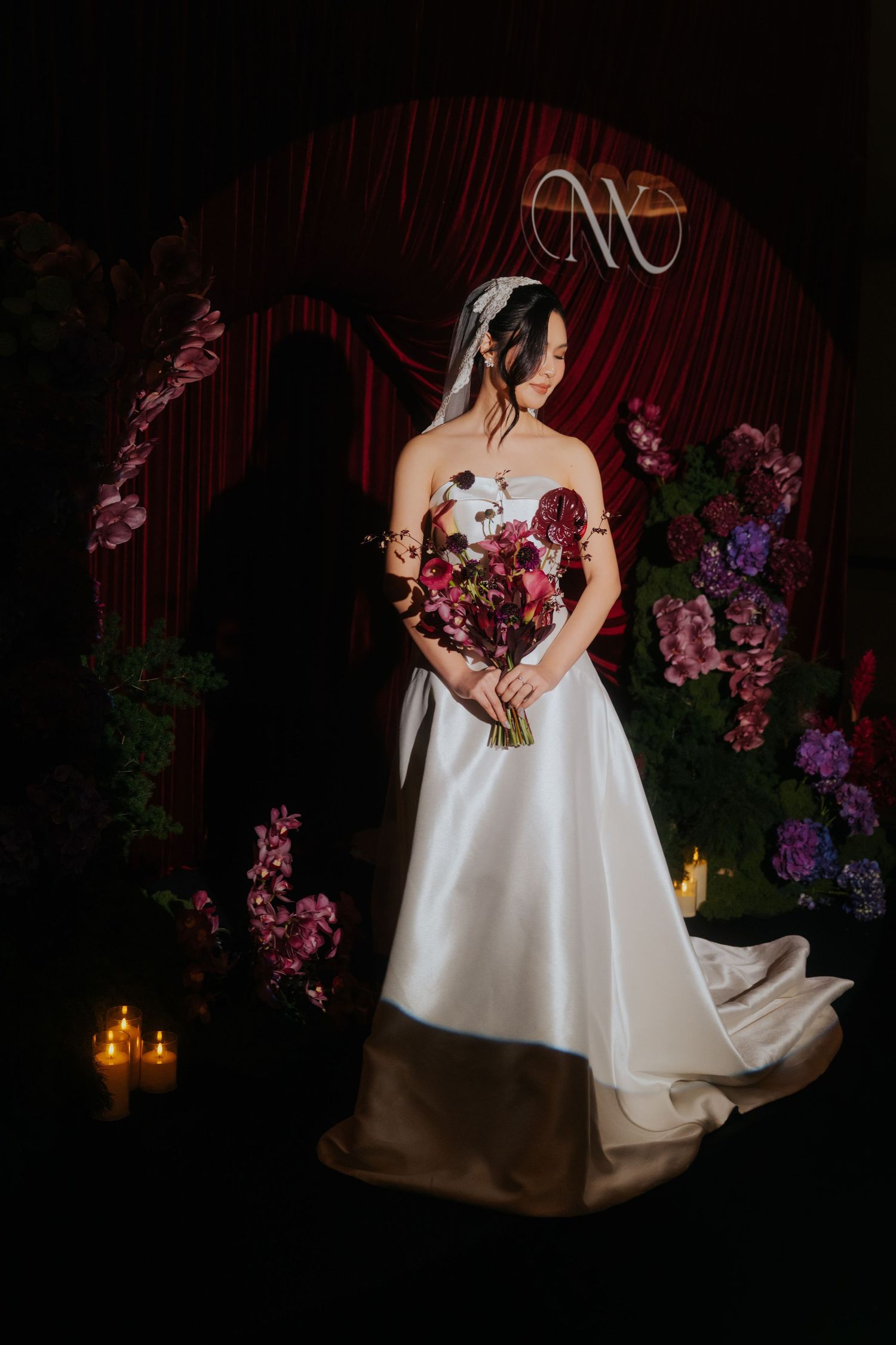 Bride in strapless white gown holds burgundy bouquet before floral arch with candles and red drapes.