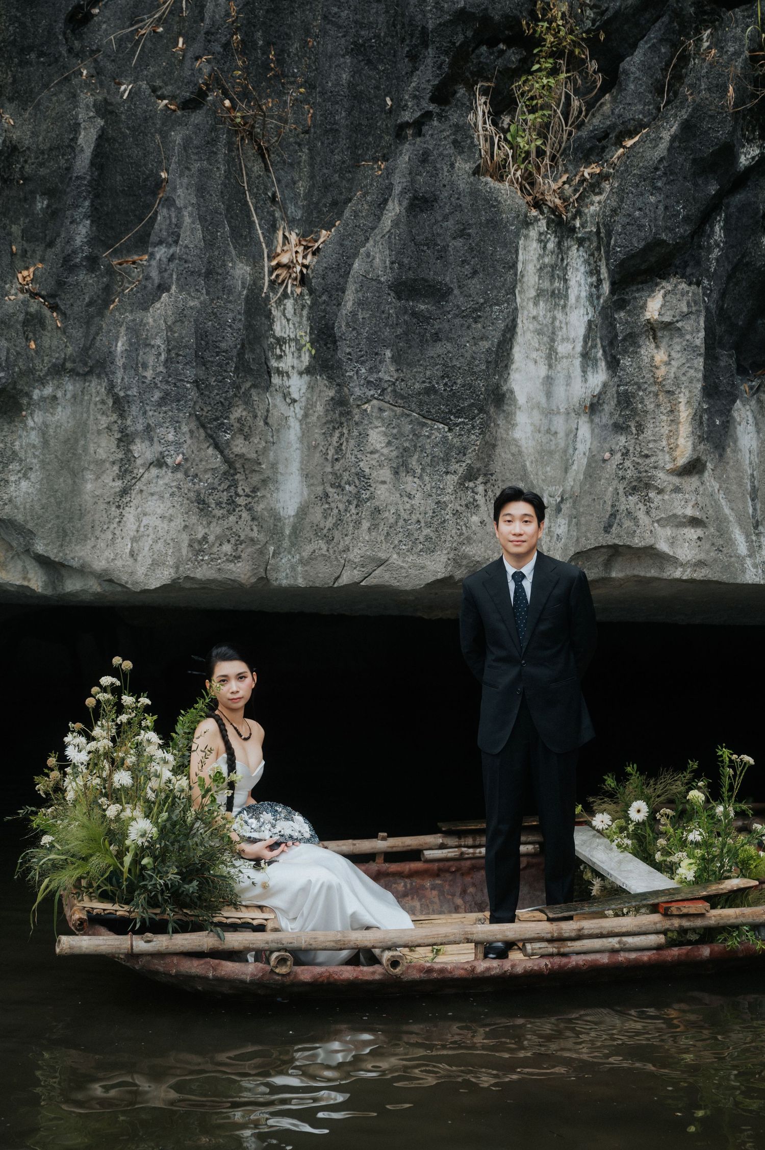 Couple in formal attire on a flower-adorned boat near dramatic rock cliffs over dark water.