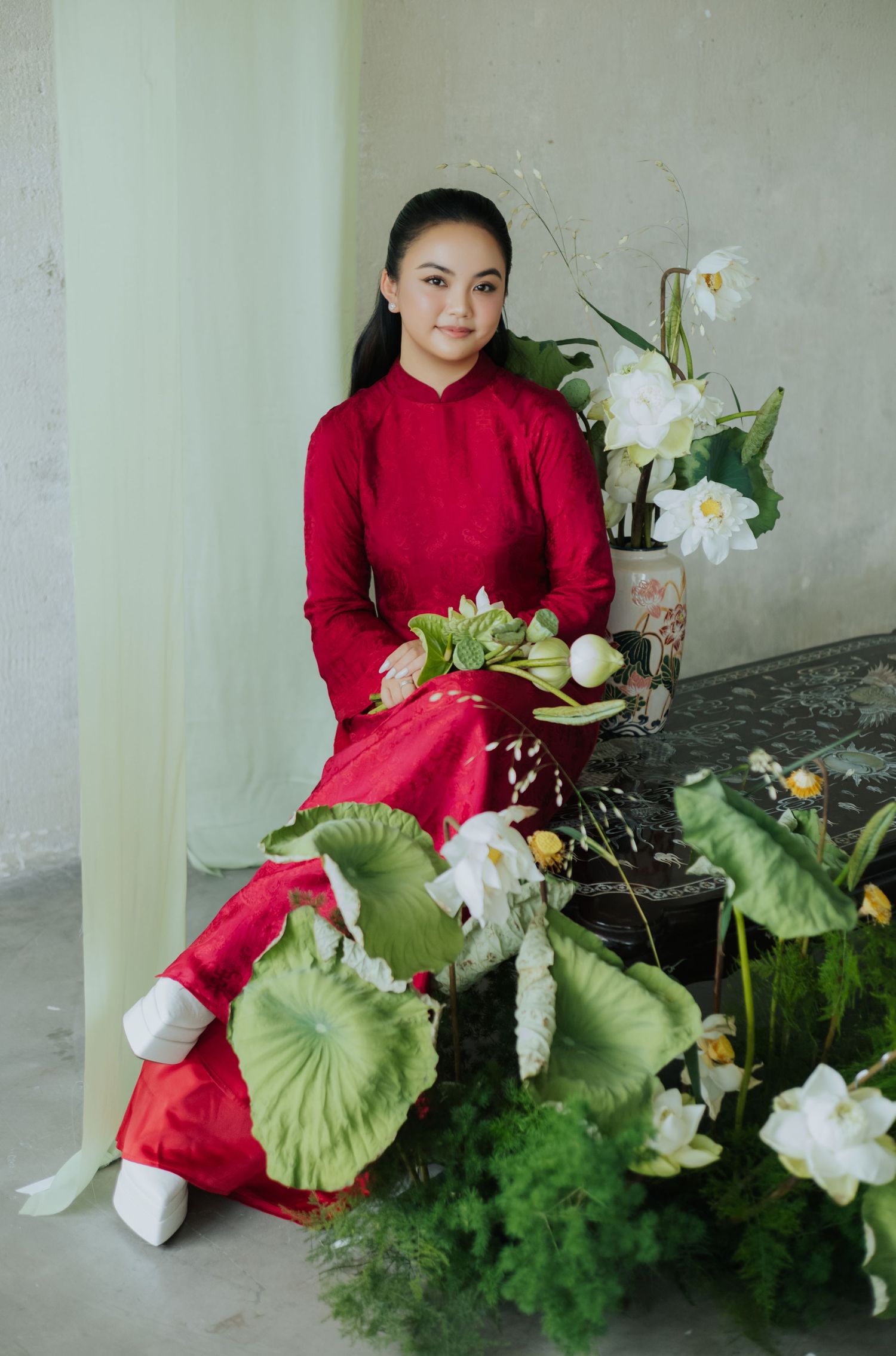 Young girl in red Vietnamese ao dai sits gracefully among white lotus flowers and green leaves in elegant setting.