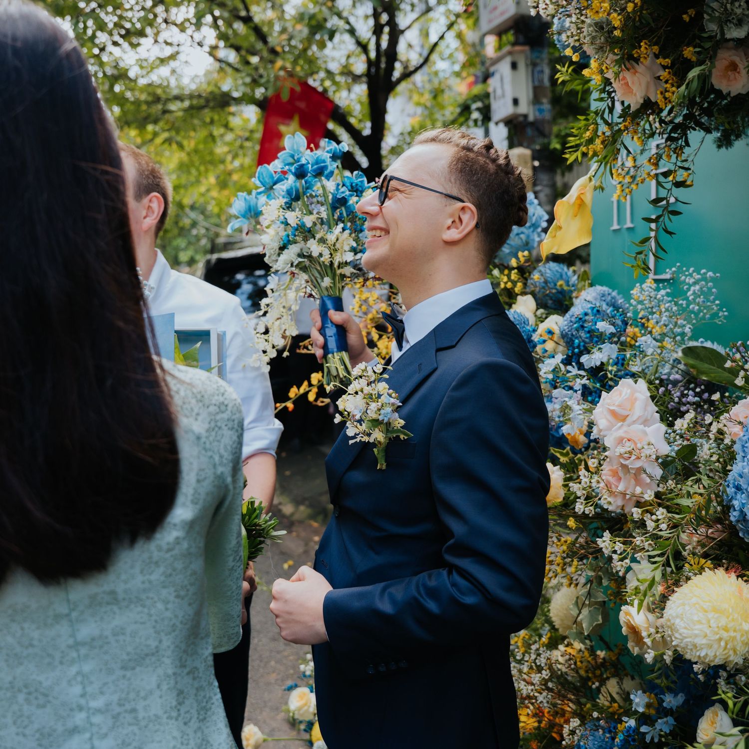 Man in navy suit smiling among colorful blue and yellow floral arrangements at an outdoor celebration.