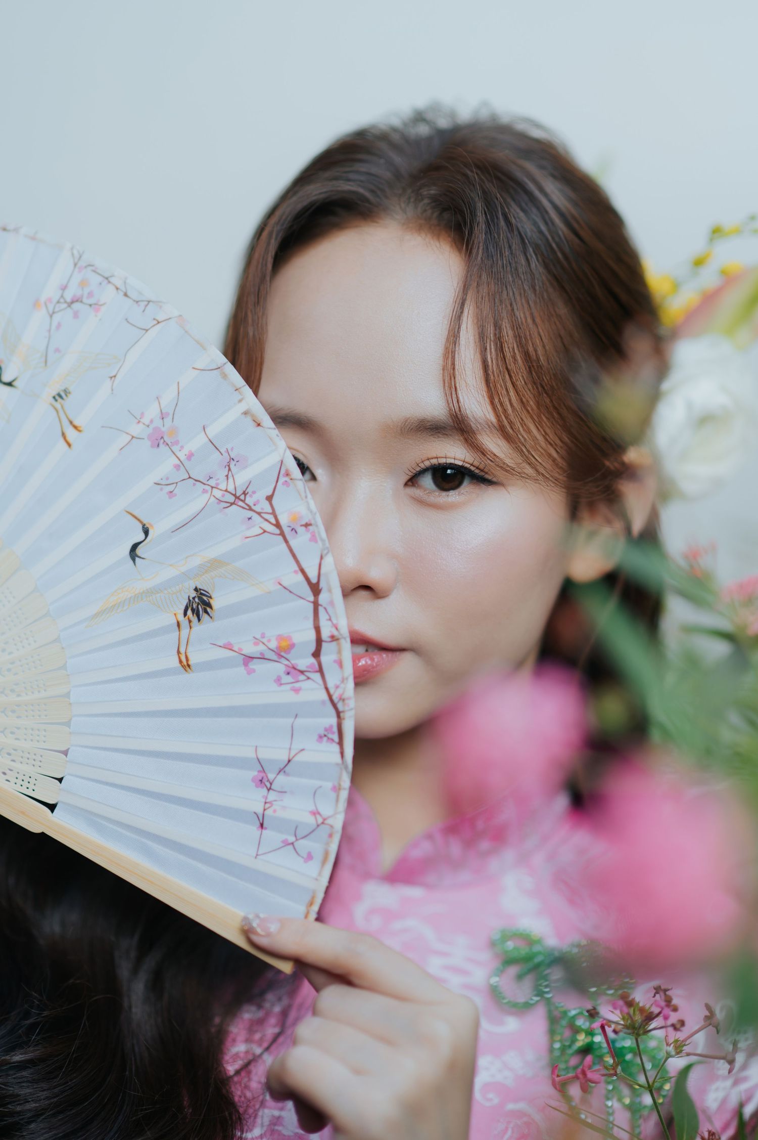 Young Asian woman in pink dress holding a decorative floral hand fan near her face with pink flowers in background.