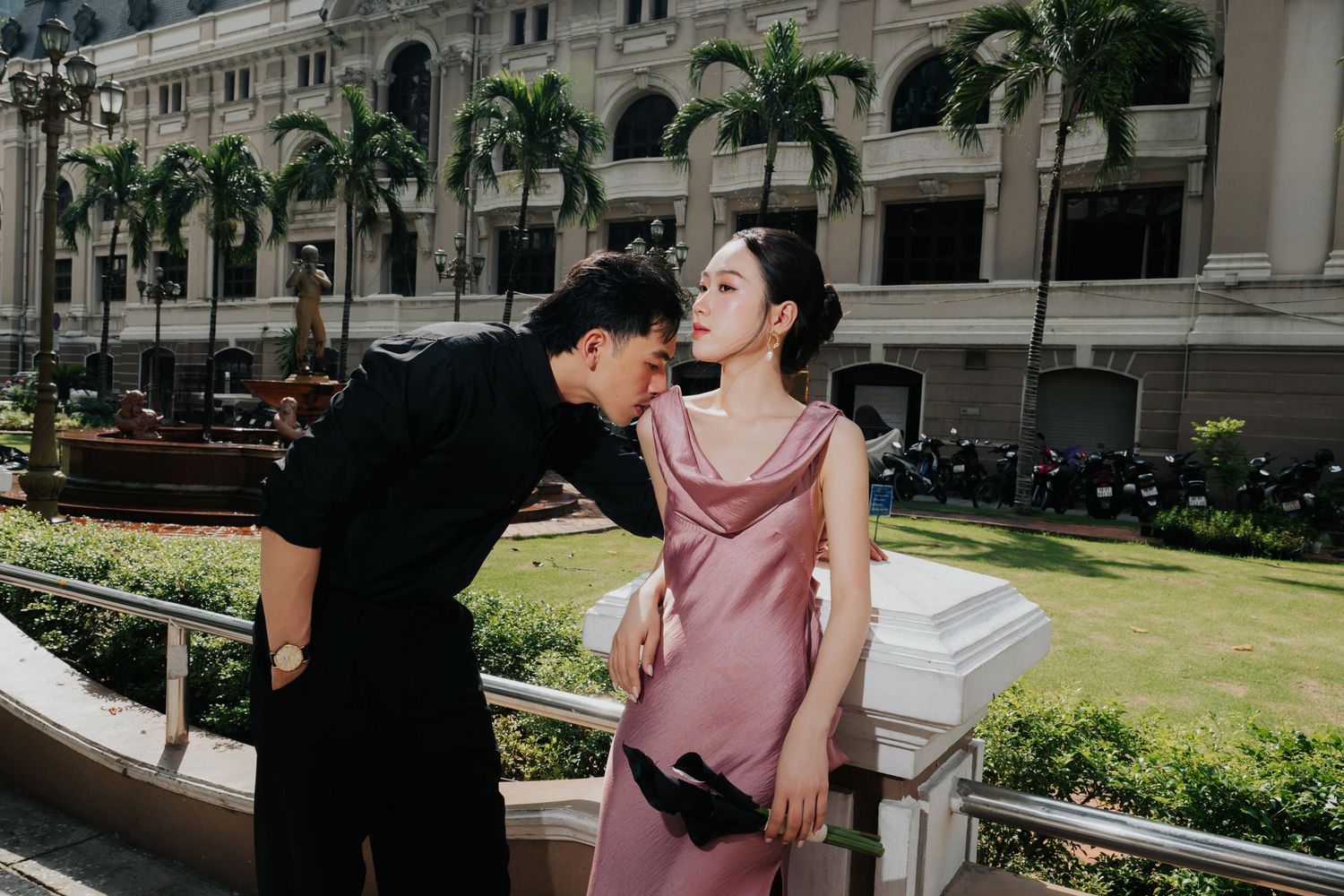A couple shares a romantic moment outside a grand colonial building, man in black kissing woman in pink satin dress.