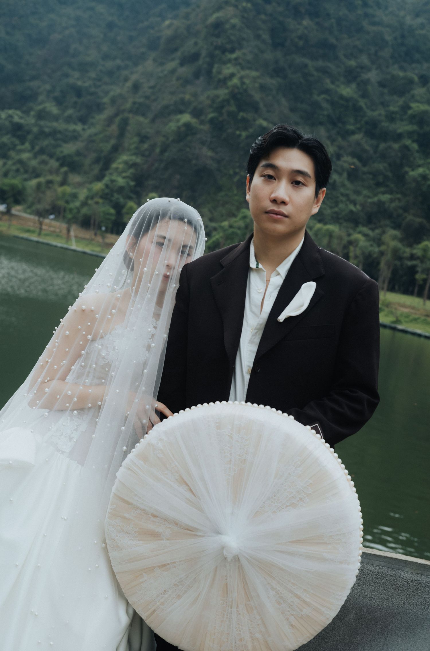 A couple poses for wedding photos by a lake, the bride in a white gown and veil holding a traditional Vietnamese hat.