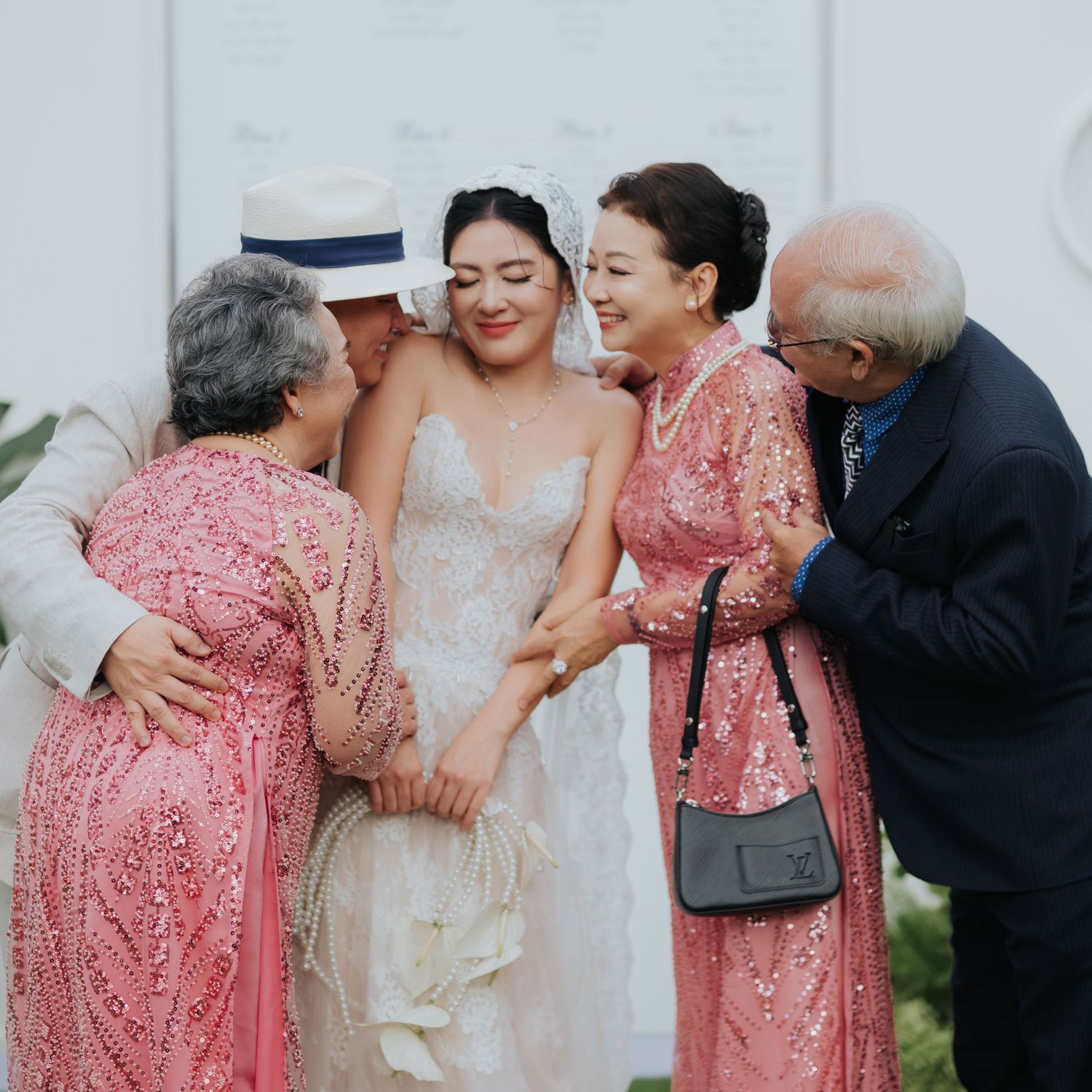 Bride in white lace gown surrounded by family in pink dresses, sharing joyful embraces at a wedding celebration.