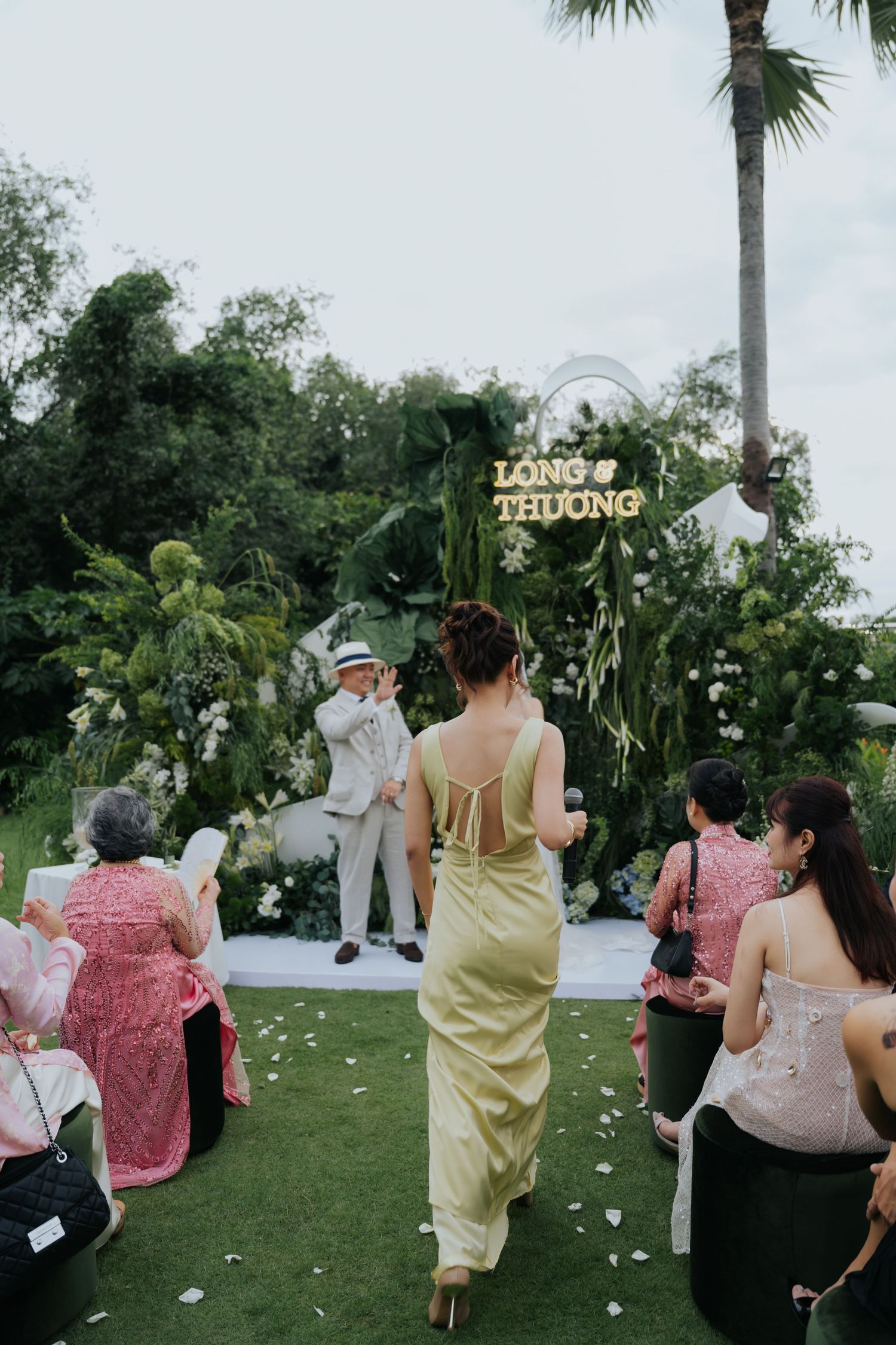 Outdoor wedding ceremony with guests seated on lawn, bride walking toward groom at floral arch reading Long & Thuong.