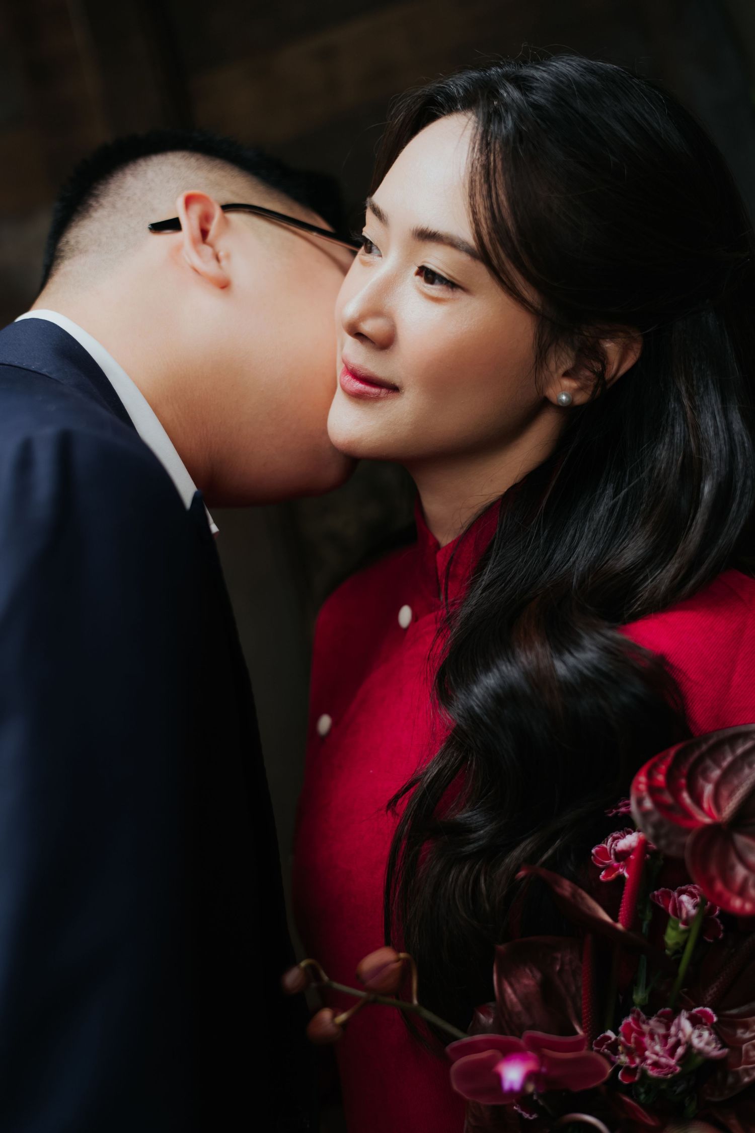 A man in a suit kisses a woman in a red dress on the cheek as she smiles holding red flowers.