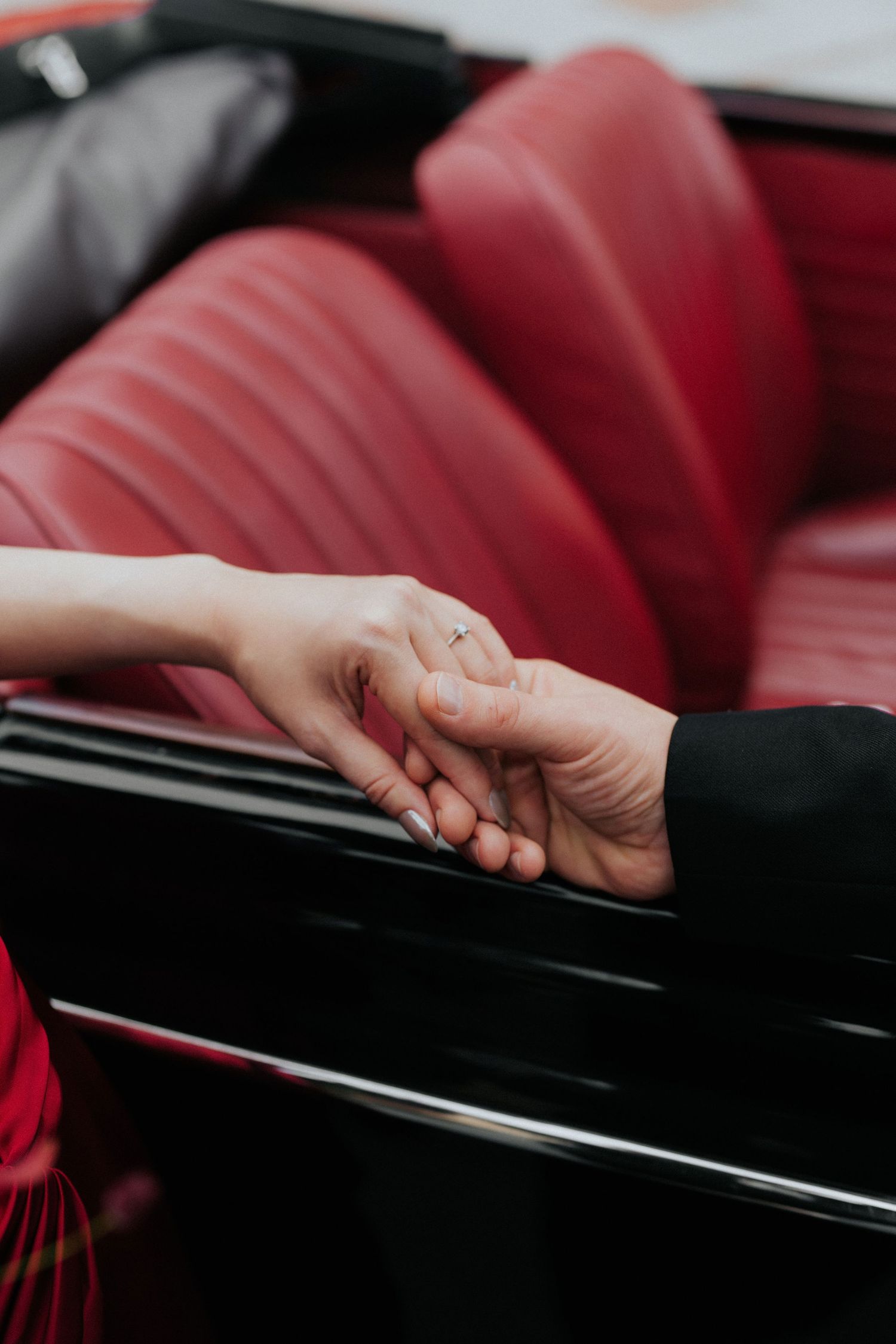 Two hands holding tenderly over a vintage car with red leather seats, woman wearing an engagement ring.
