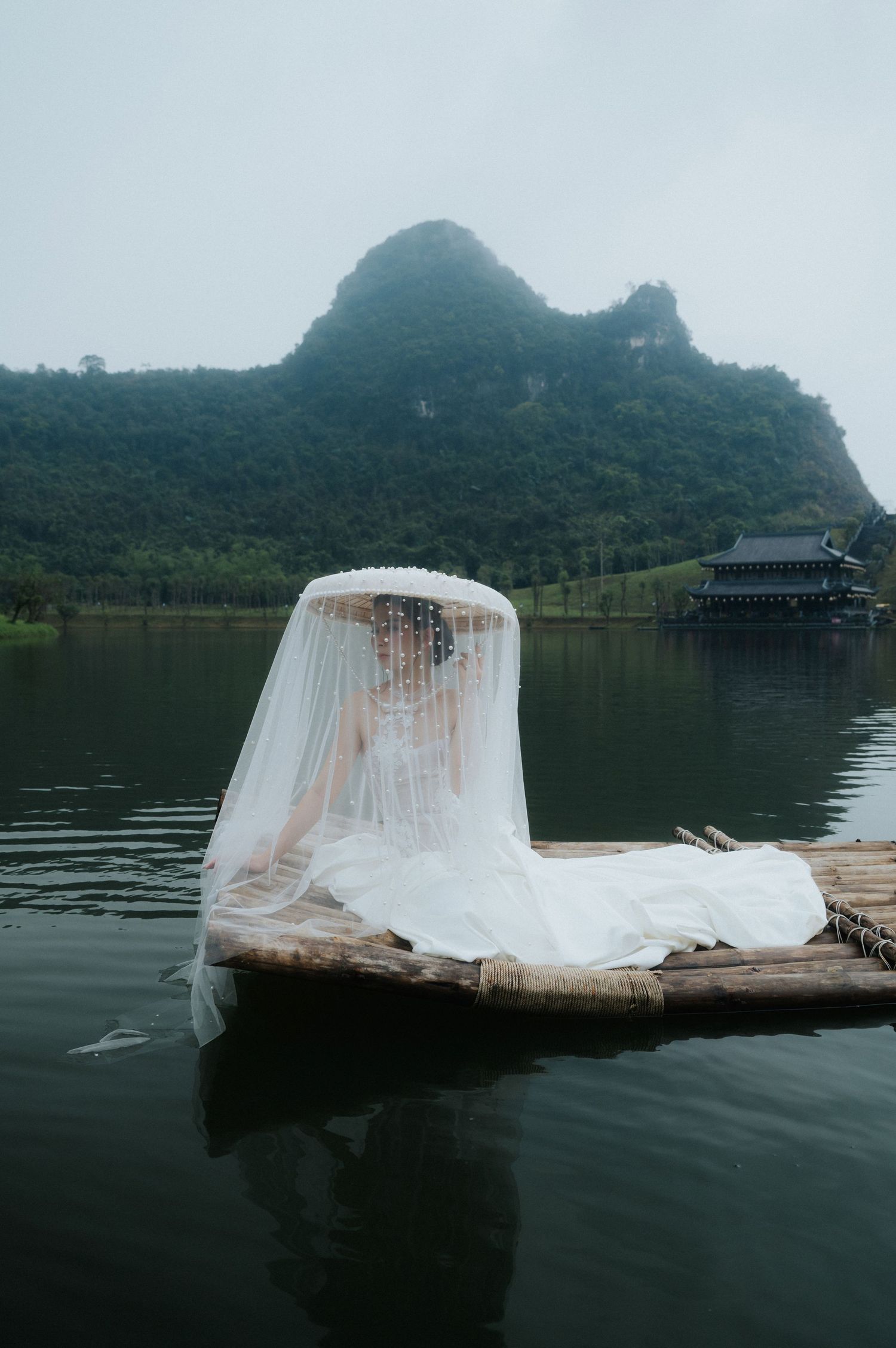 Bride in white wedding dress and veil sits on bamboo raft on calm lake with misty karst mountains in background.