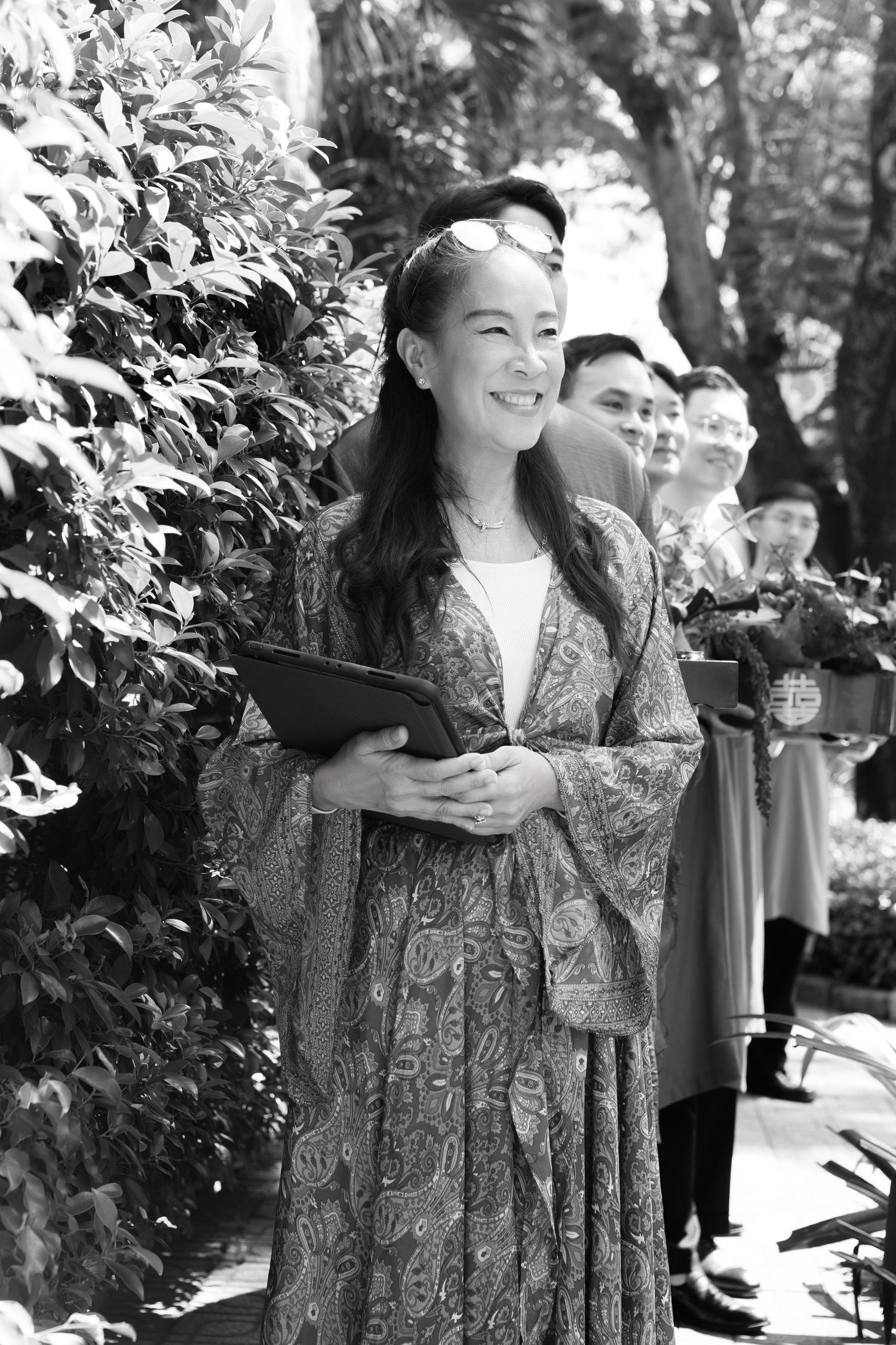 Woman in patterned kimono-style dress smiling outdoors, holding a tablet, with greenery and people in background.