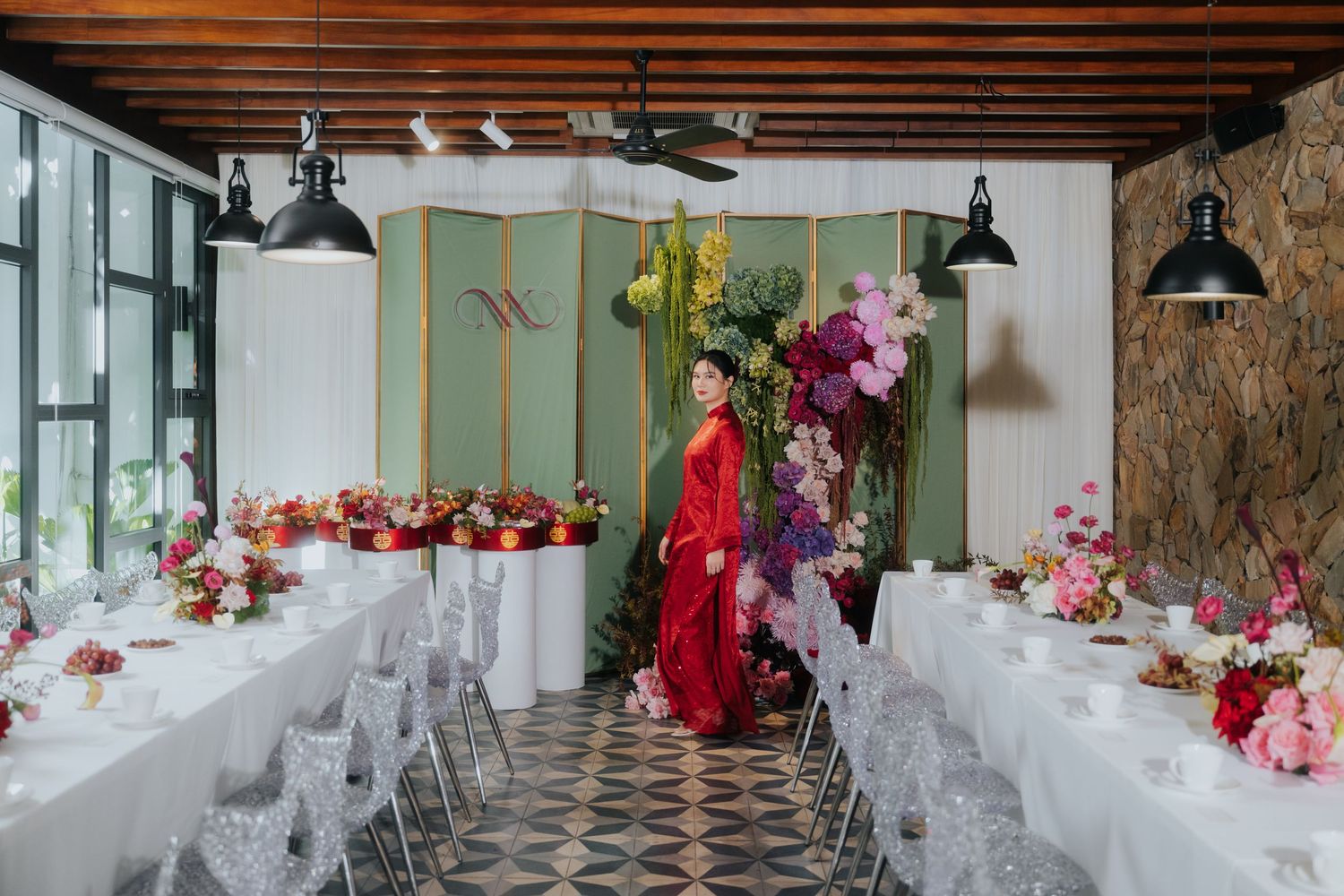 A woman in a red gown stands among floral arrangements in an elegant wedding reception venue with white tables.