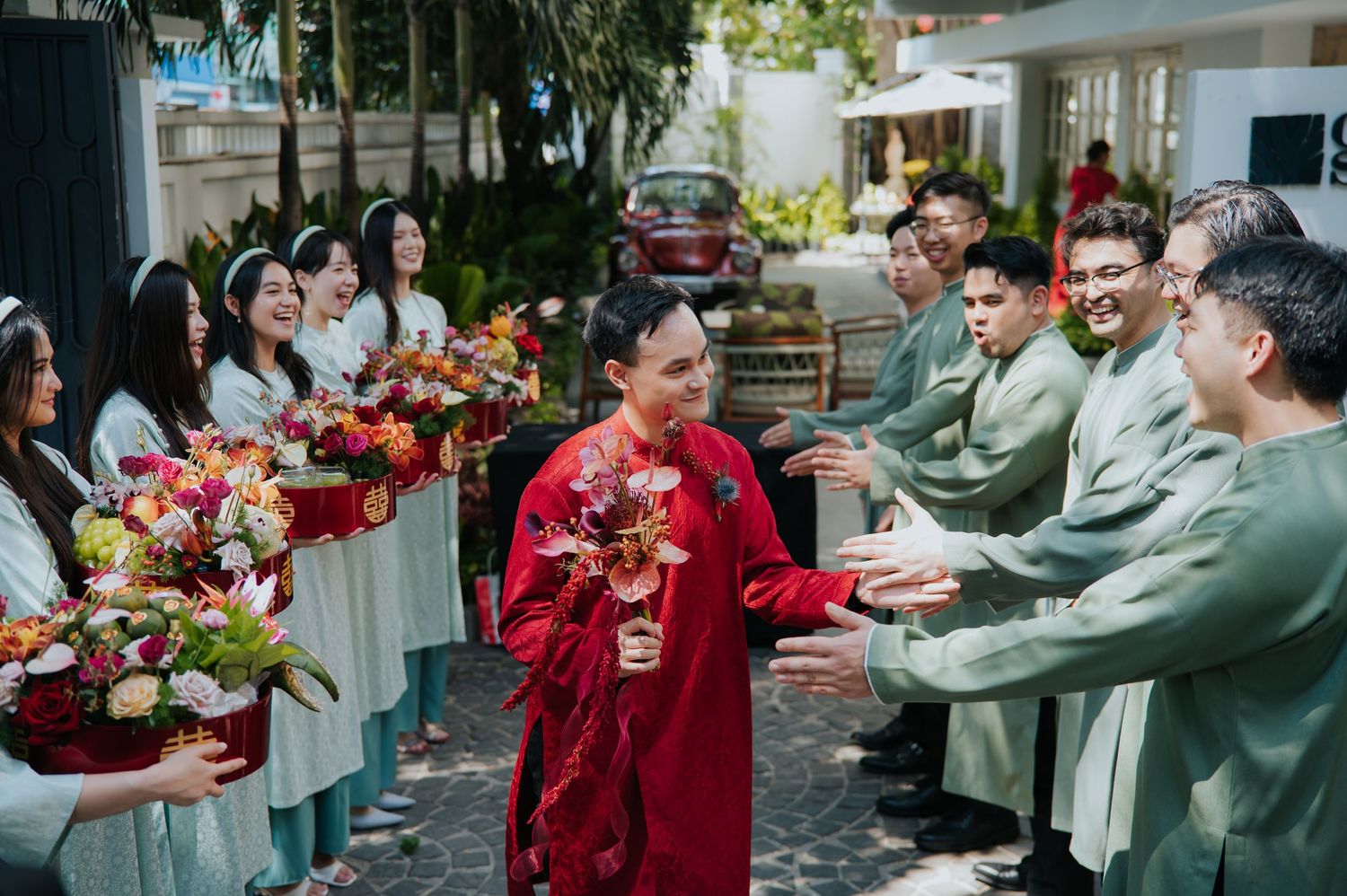 Vietnamese wedding ceremony with groom in red ao dai greeting groomsmen in green, bridesmaids in white ao dai holding floral bouquets.