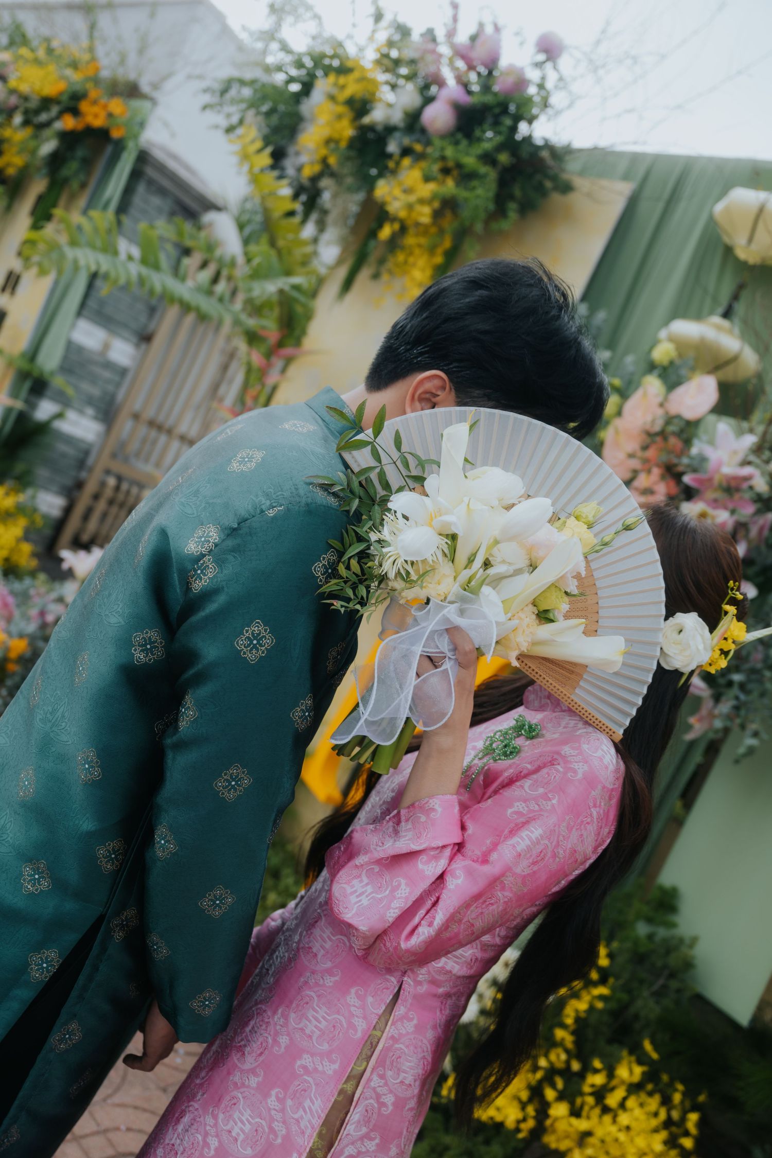 Vietnamese couple in traditional ao dai attire sharing a kiss behind a white fan and floral bouquet at a wedding ceremony.