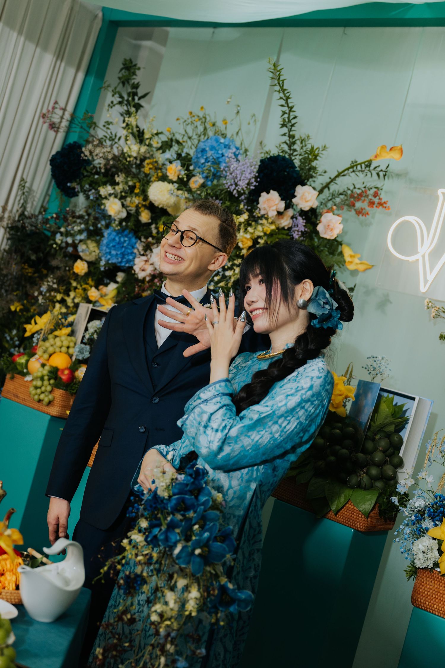 A couple poses happily in front of a colorful floral backdrop, the woman in a blue dress and man in a dark suit.