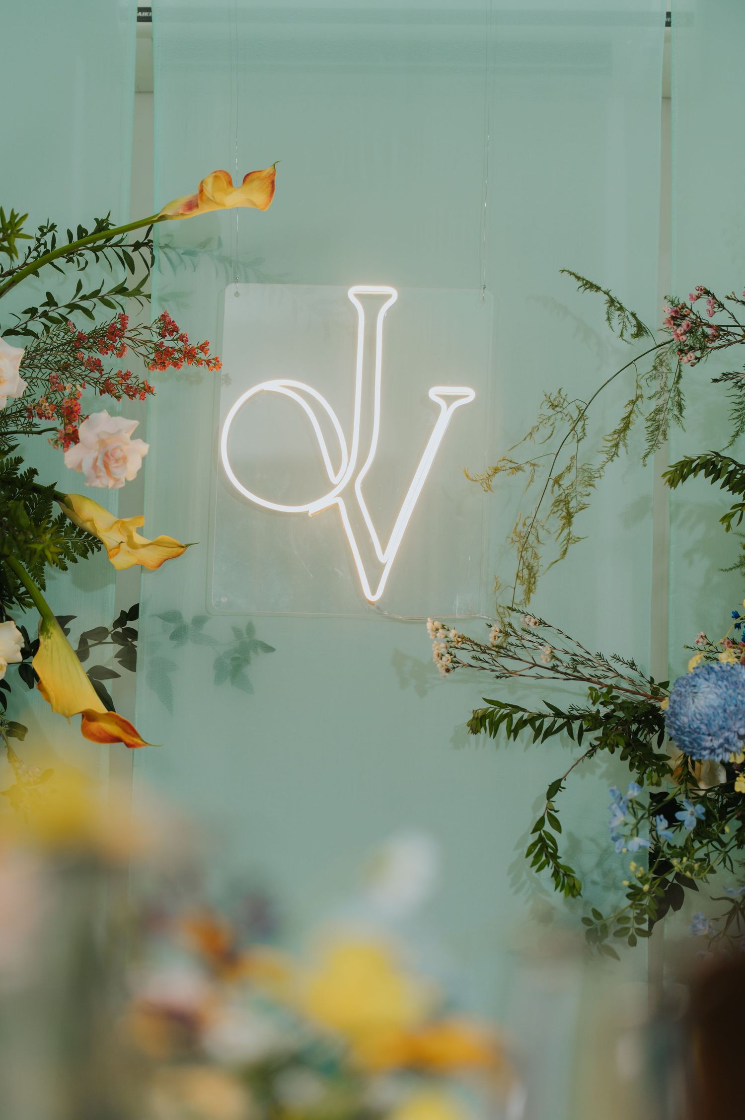 Neon monogram sign with letters DV on mint green wall surrounded by colorful floral arrangements at a wedding reception.