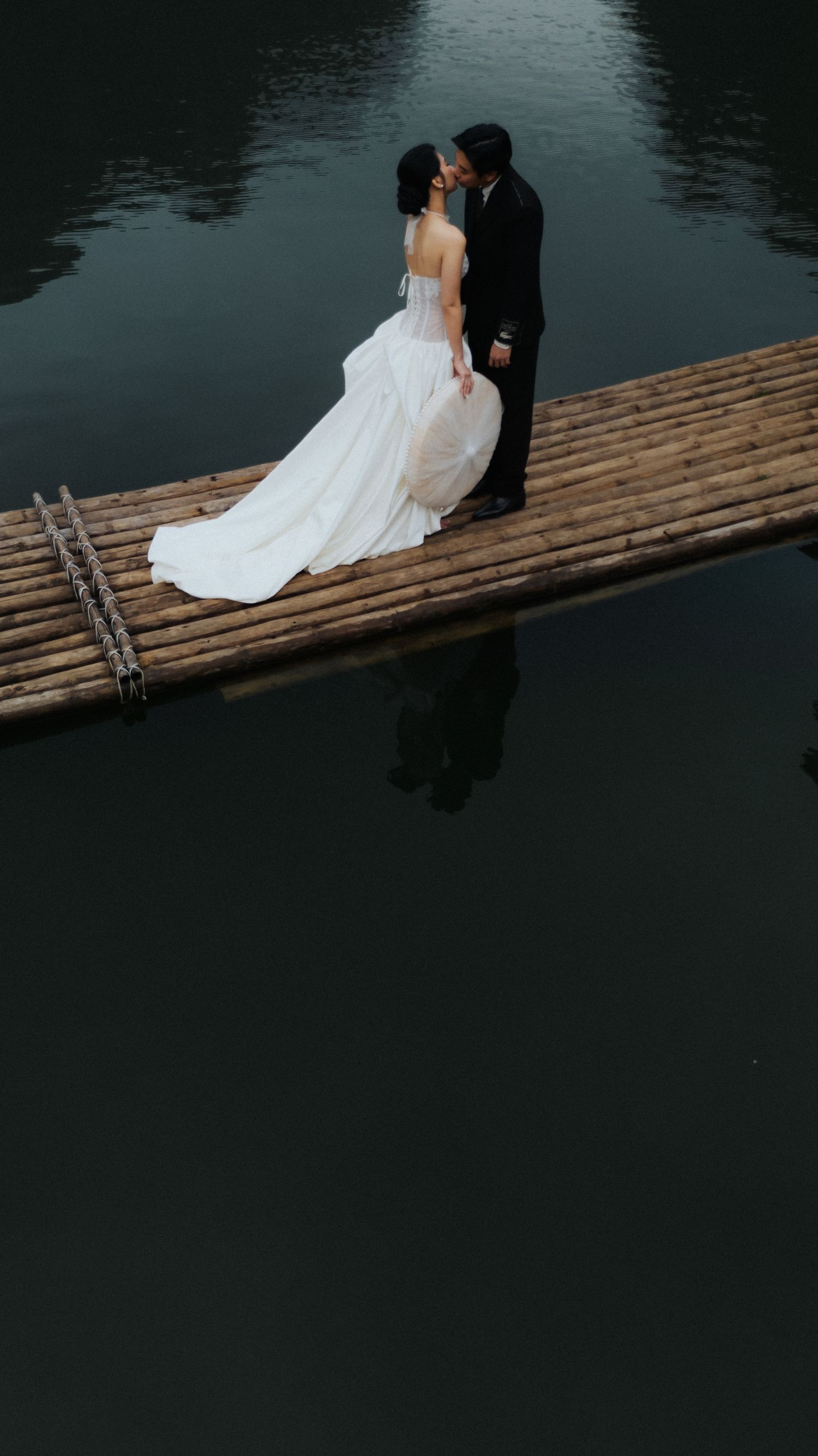 A bride and groom share a romantic kiss on a bamboo raft over calm dark water in a stunning wedding photo.