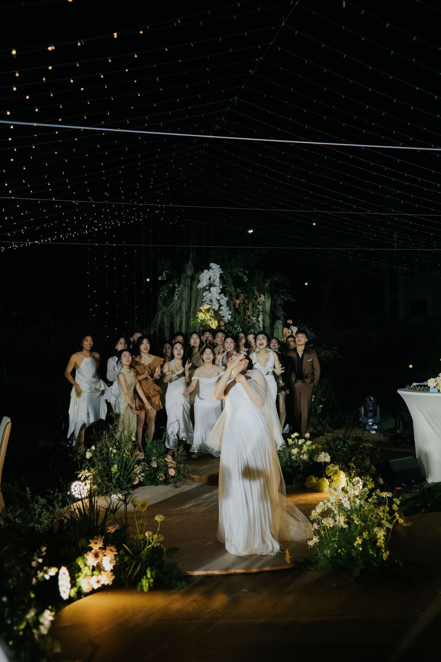 Bride tossing bouquet to guests at an elegant outdoor wedding reception with fairy lights and lush floral arrangements.