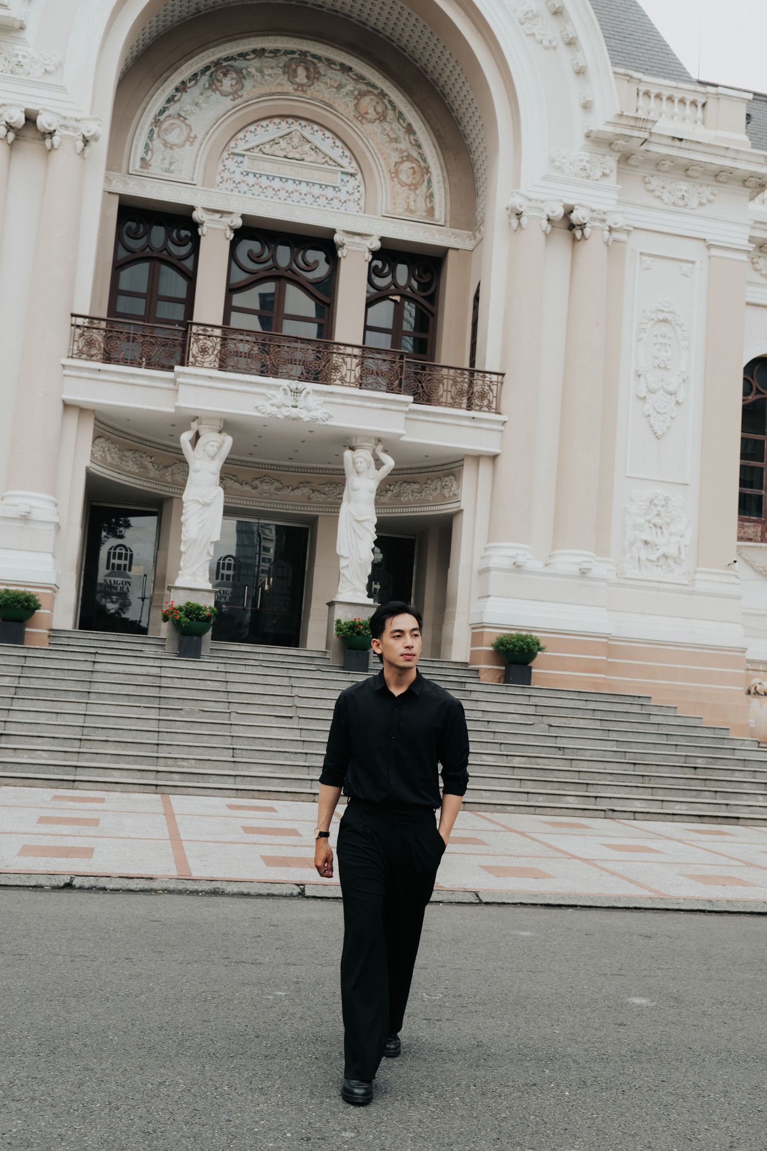Young Asian man in all-black outfit standing before a grand white neoclassical building with ornate columns.