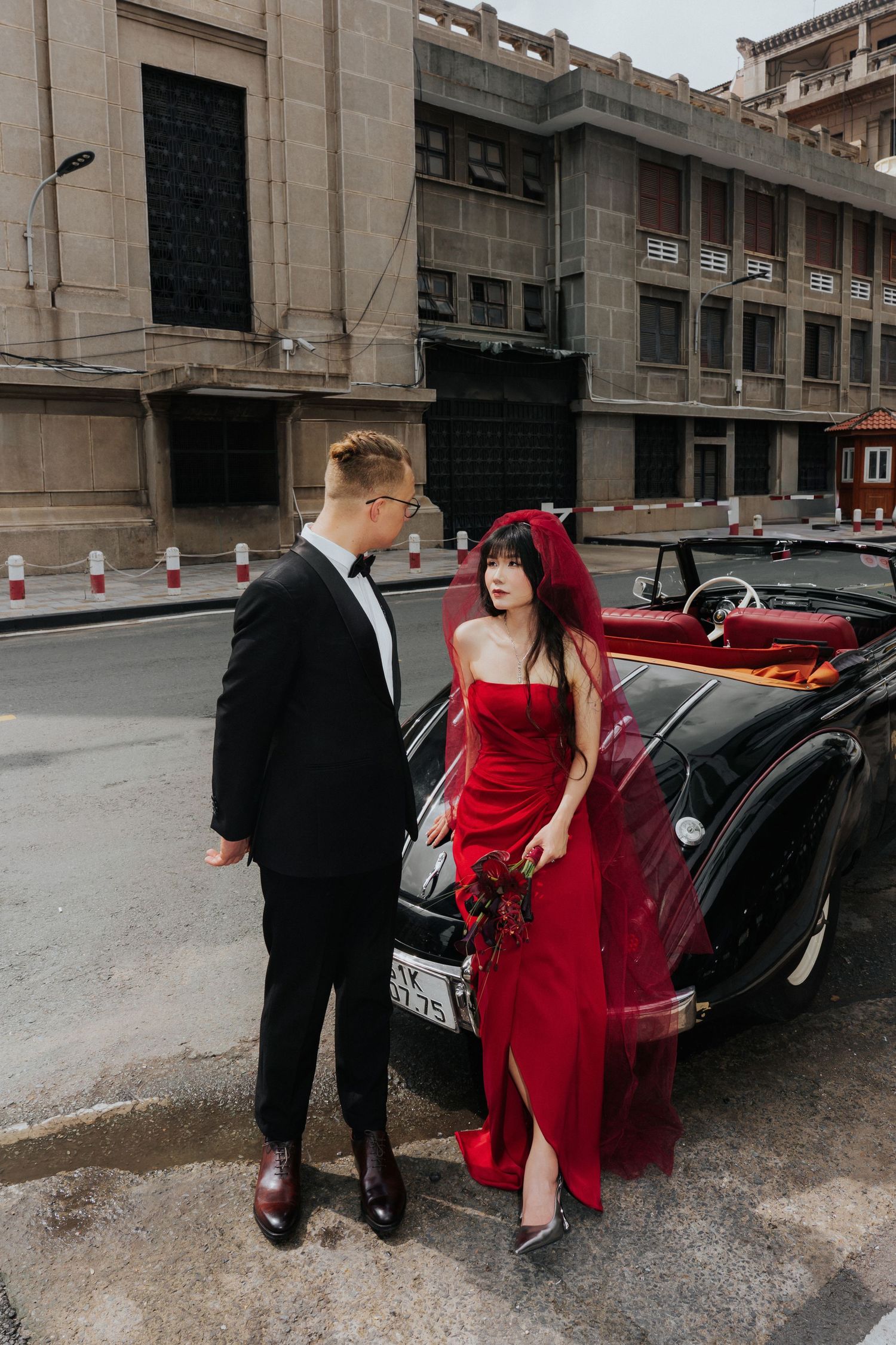 Couple in elegant wedding attire beside a vintage black convertible, bride in red gown, groom in black tuxedo, urban backdrop.