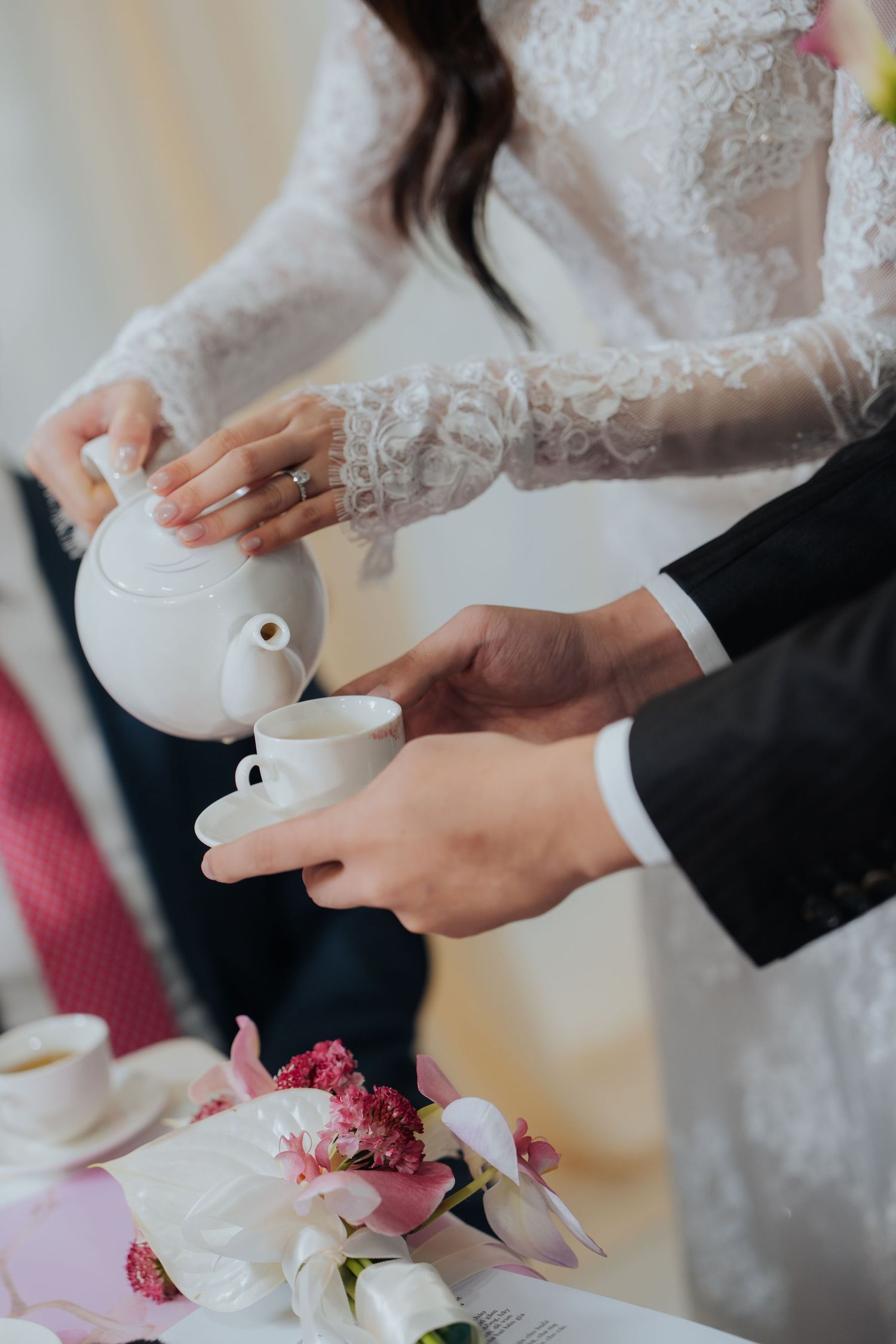 Bride in lace dress pouring tea from white teapot into cup held by groom in black suit at wedding ceremony.