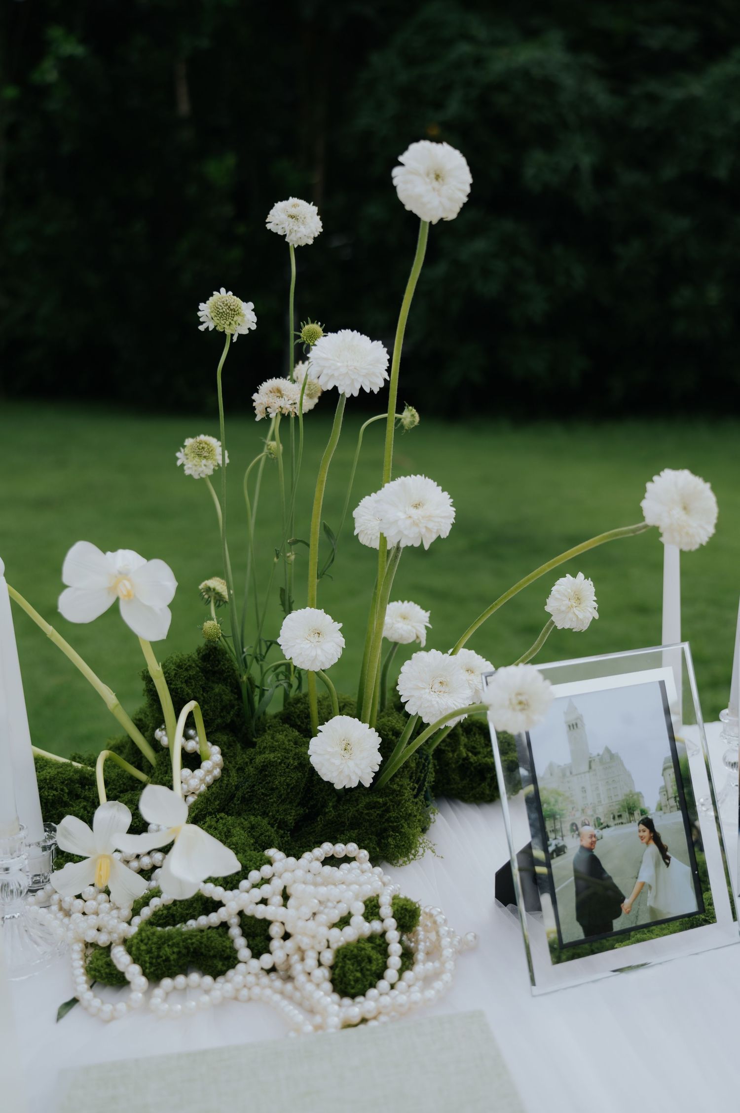 White ranunculus and orchids arranged on moss with pearl necklaces and a framed wedding photo on a white table outdoors.