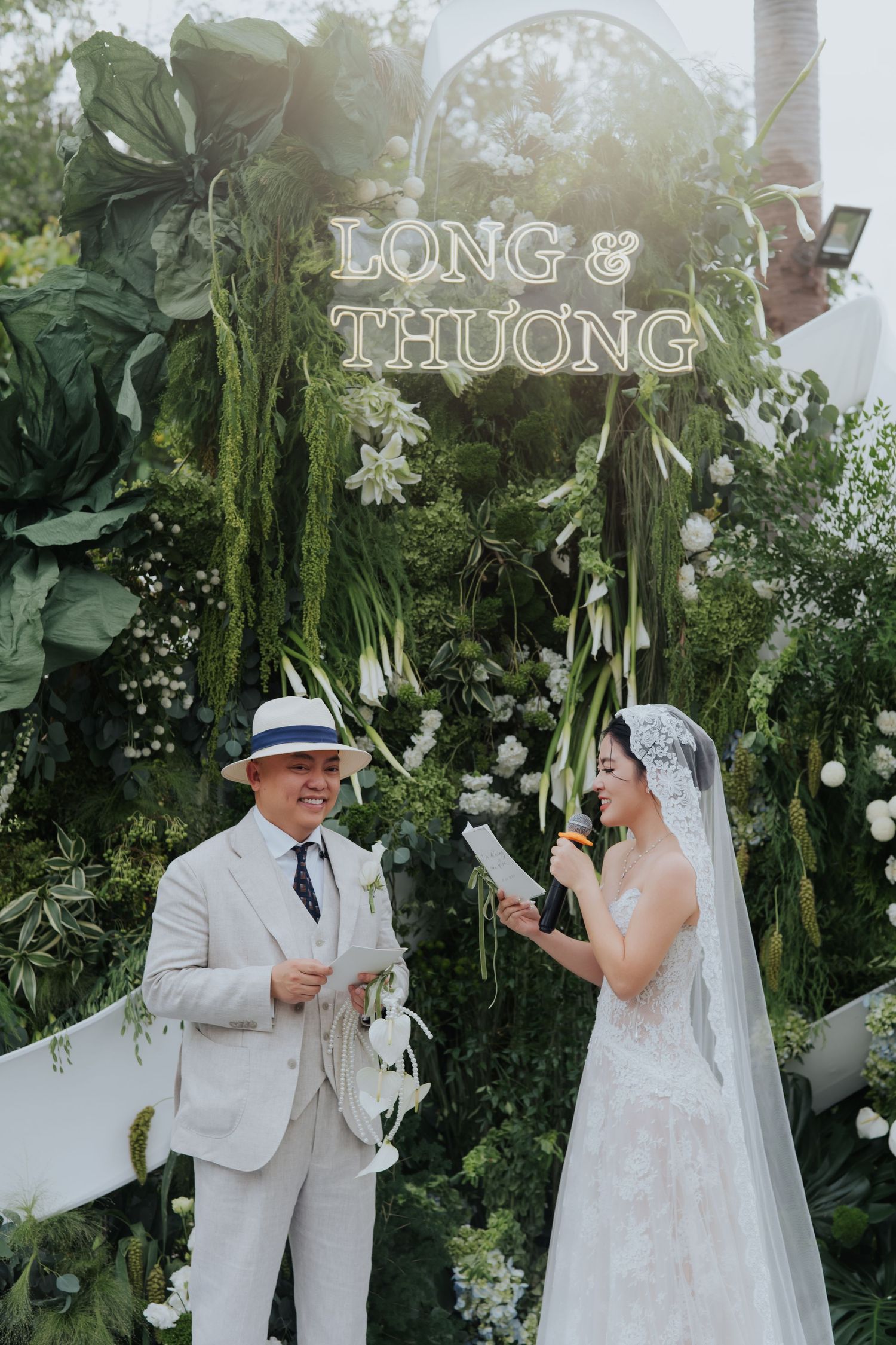 Couple exchanges vows at outdoor wedding ceremony beneath lush green floral arch with Long & Thuong neon sign.
