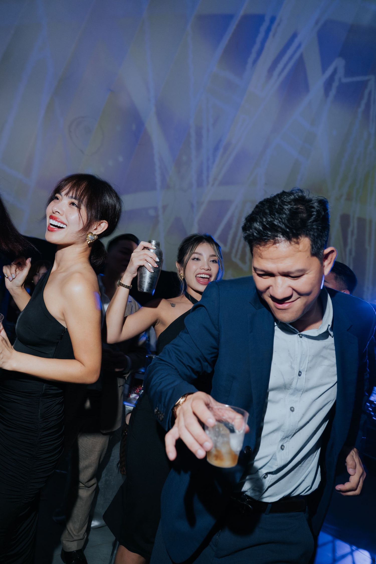 Young adults dancing and celebrating at a party, holding drinks and smiling under blue event lighting.