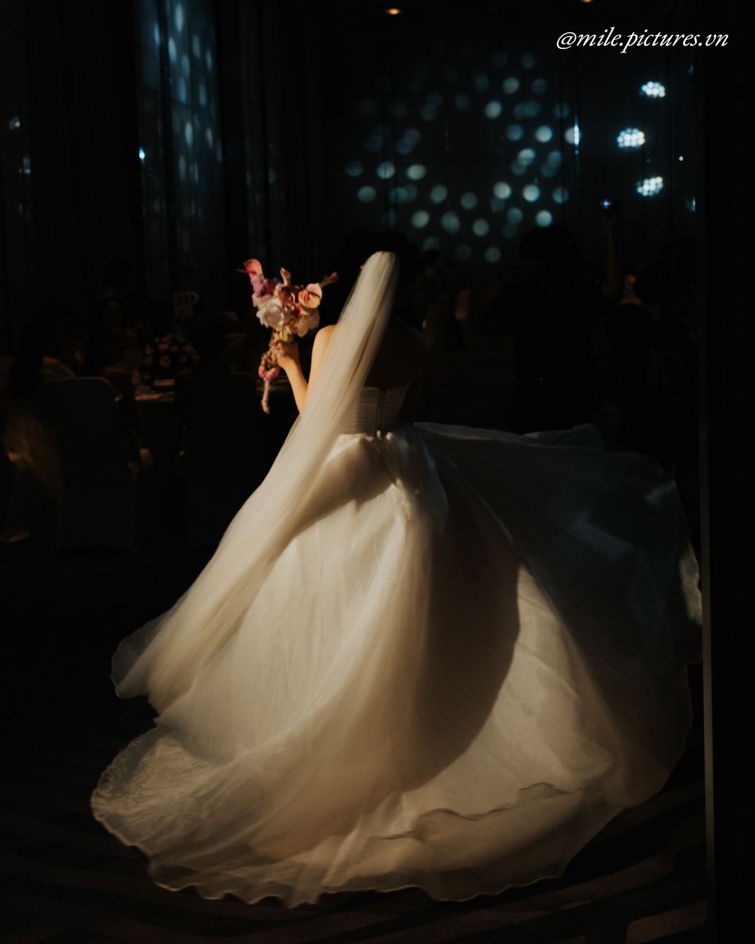 Bride in flowing white wedding gown twirling gracefully in dramatic dark venue lighting, holding colorful bouquet.