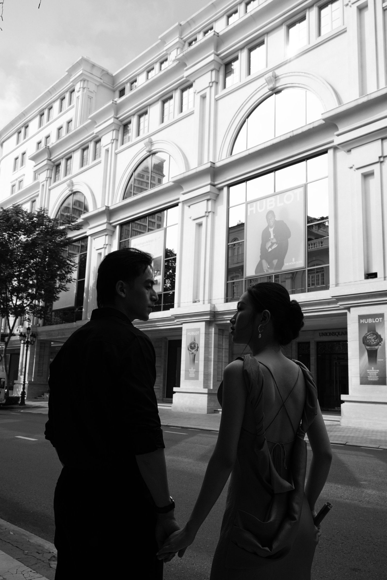 Black and white photo of a couple holding hands in front of a grand classical building on a city street.