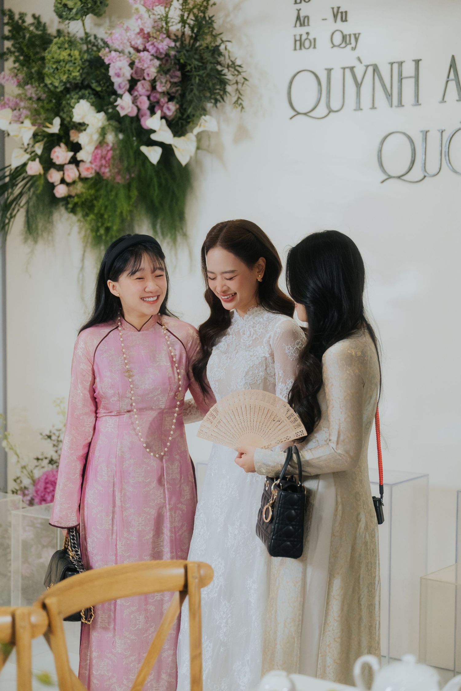 Three women in elegant ao dai attire smile together at a Vietnamese engagement ceremony decorated with flowers.