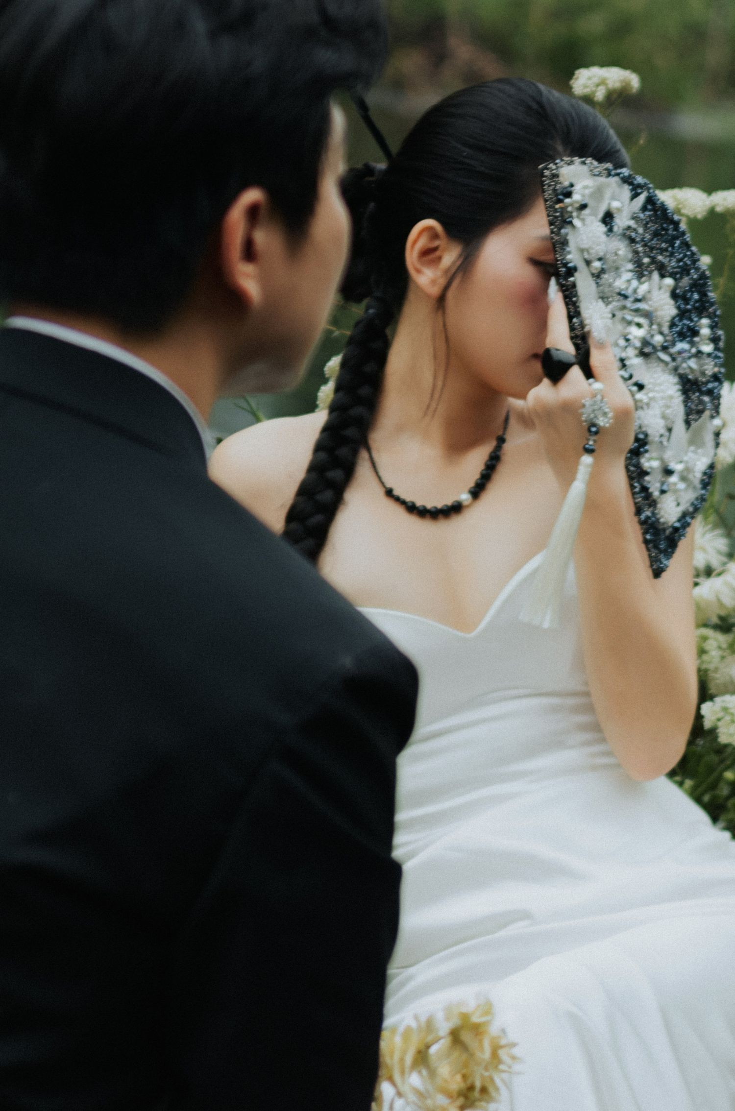 A bride in a white gown and black beaded necklace holds a floral bouquet beside a groom in a black suit.