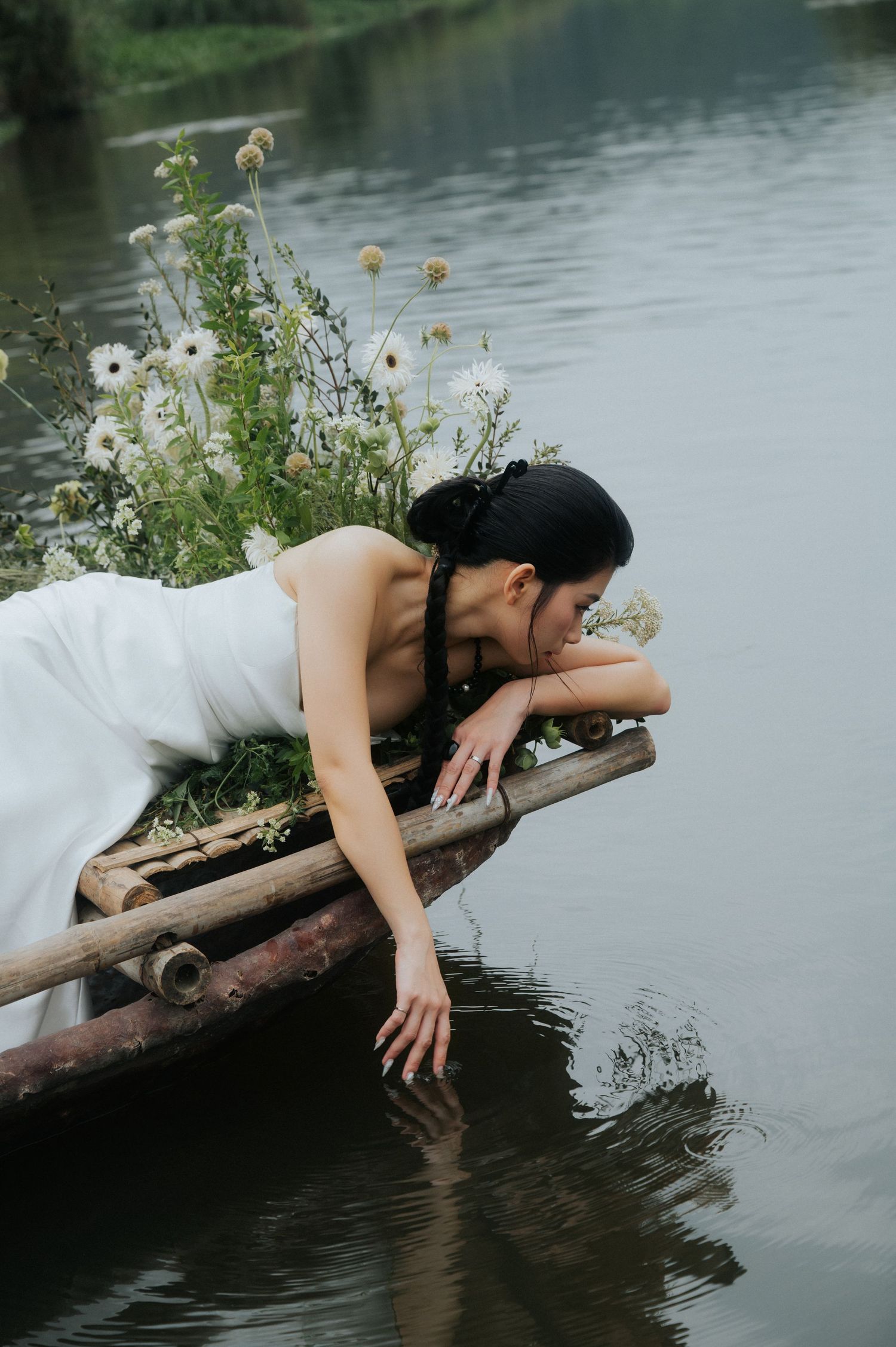 A woman in a white dress leans over a bamboo raft, touching the calm water, surrounded by wildflowers.