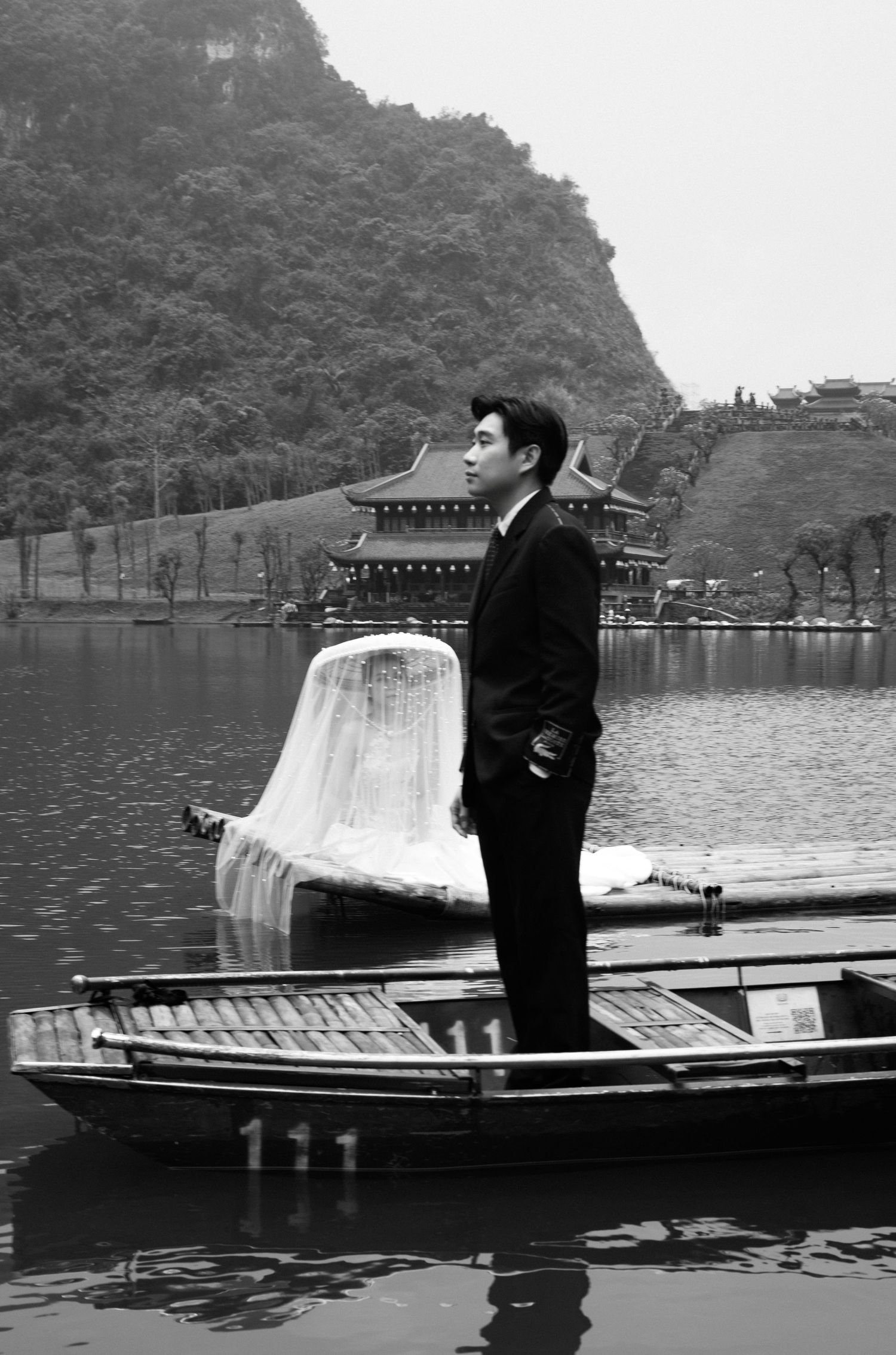 Black and white photo of a man in a suit standing on a wooden boat by a lake with an Asian temple in the background.
