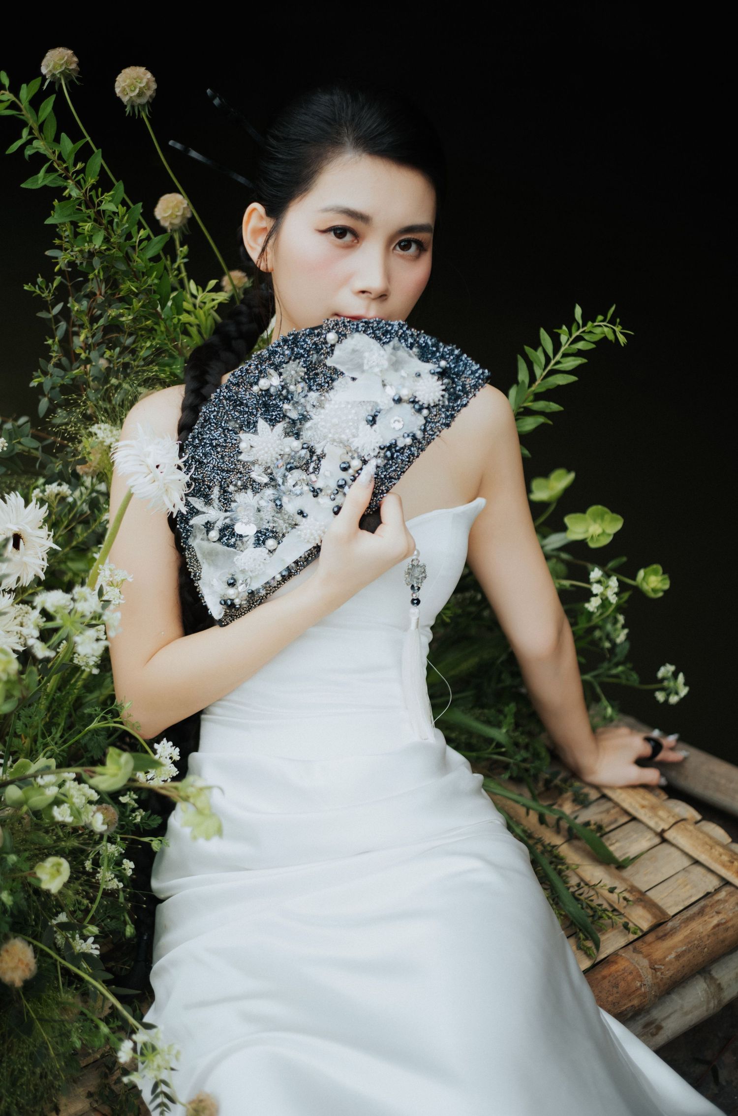Young woman in white wedding dress holding decorative fan surrounded by white flowers and green foliage.