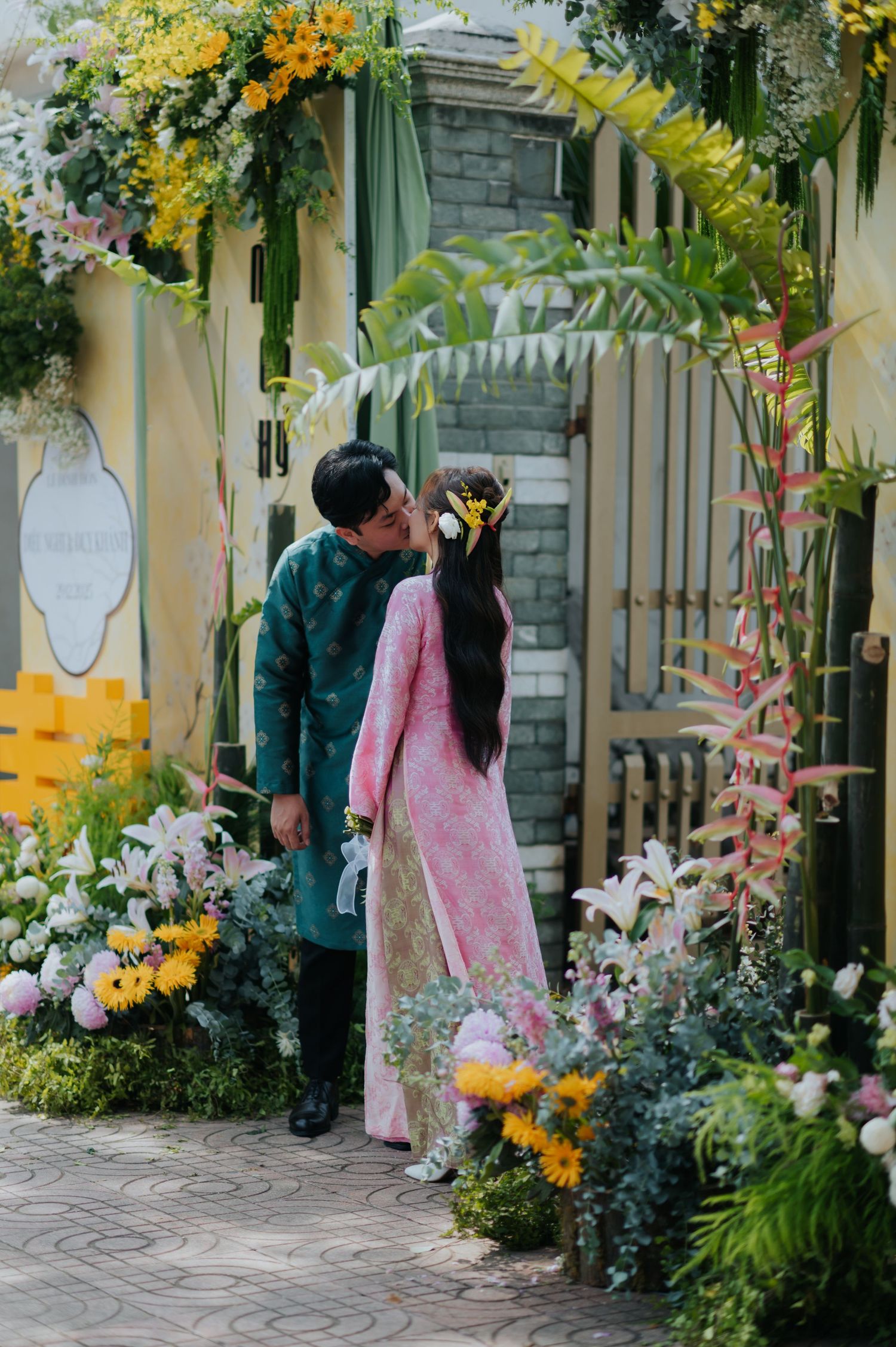 Couple in traditional Vietnamese ao dai kissing under a floral arch decorated with colorful blooms.