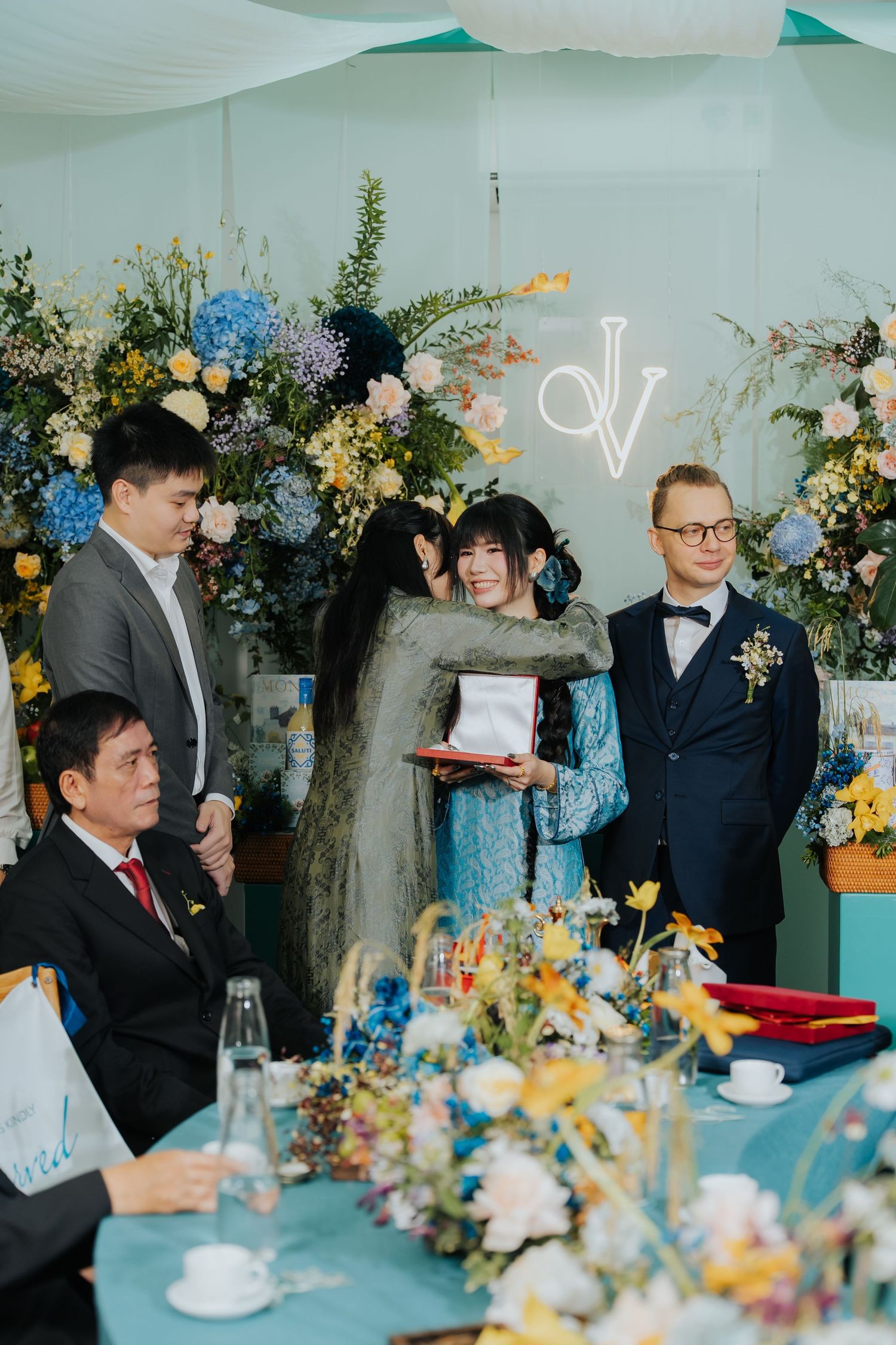 Elegant wedding ceremony with guests exchanging gifts near a floral backdrop with blue hydrangeas and a monogram sign.